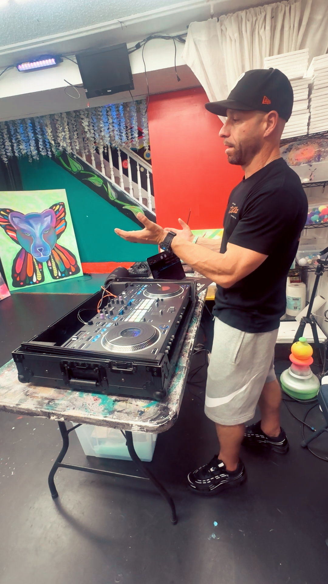 Man in black cap and black shirt stands at DJ equipment, gesturing with his hands, in a room with colorful wall art.
