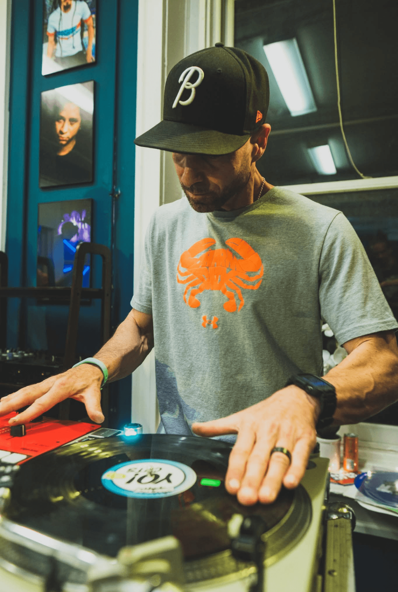 Man wearing a black cap with a white logo, gray t-shirt with orange crab, using a turntable in a dimly lit room.