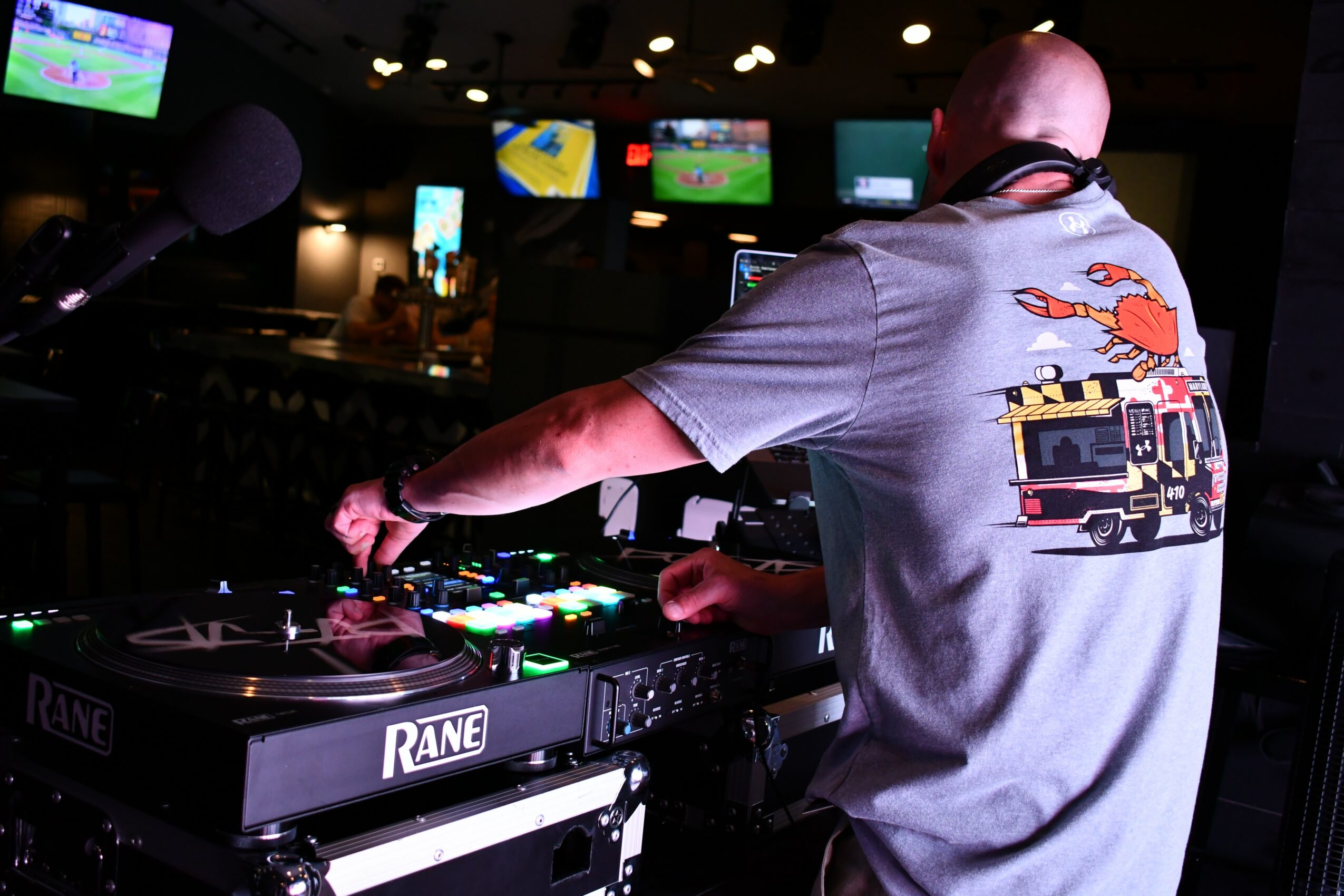 DJ wearing a gray T-shirt with a fire truck graphic, operating DJ equipment in a dimly lit venue with screens in background.