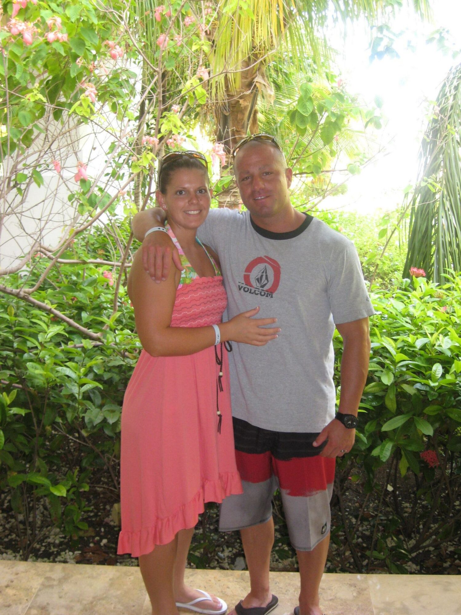 A woman and man stand together outdoors, smiling, surrounded by green plants and trees.