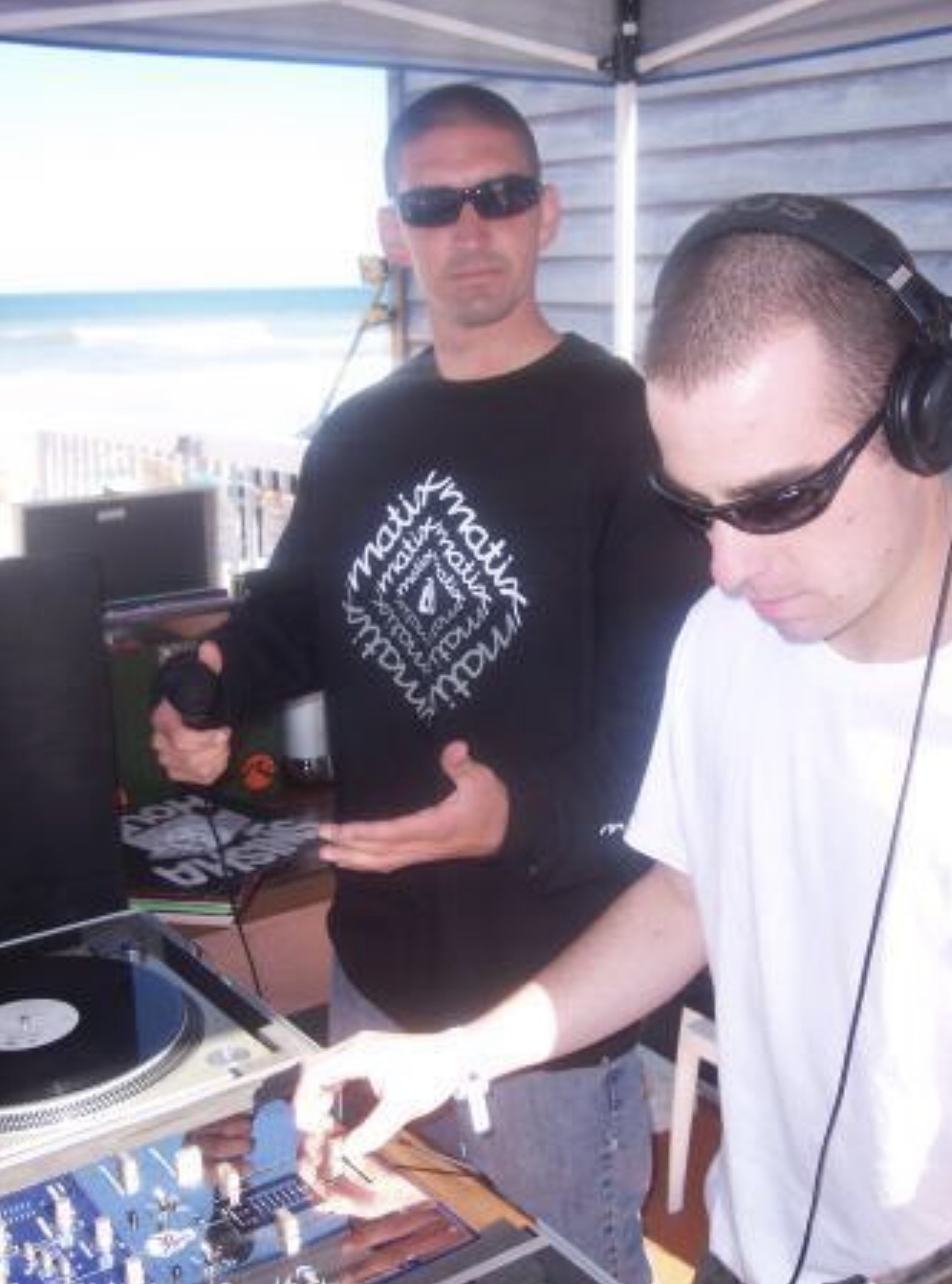 Two men working with DJ equipment outdoors, one wearing sunglasses and a black sweatshirt, the other in a white shirt and headphones.