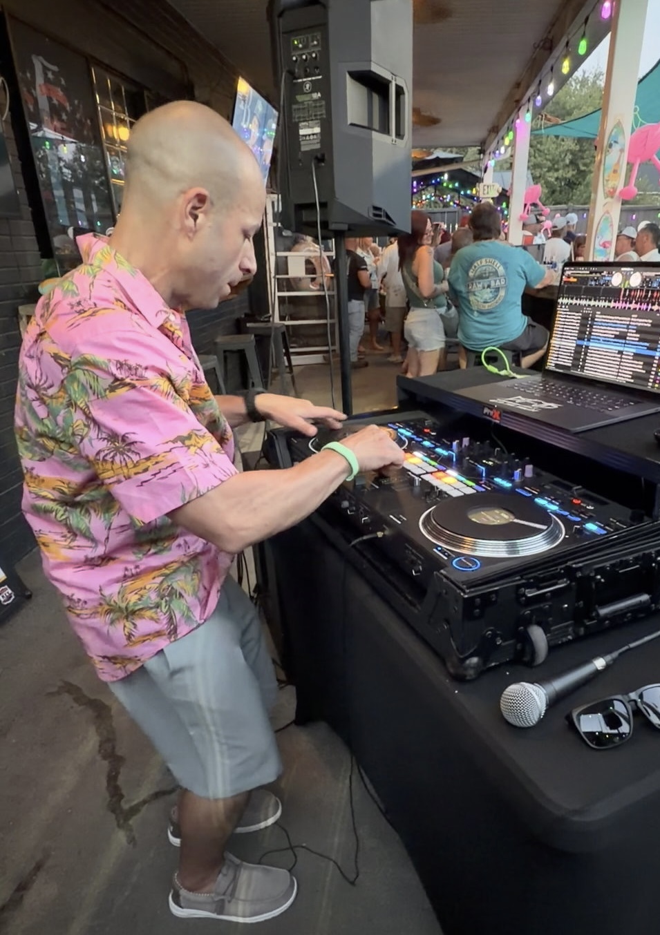 Bald man in pink floral shirt and shorts DJing at an outdoor event with people and colorful decorations.