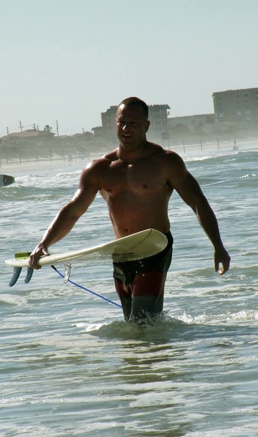 Man walking in ocean water holding a surfboard, with buildings and a beach in the background