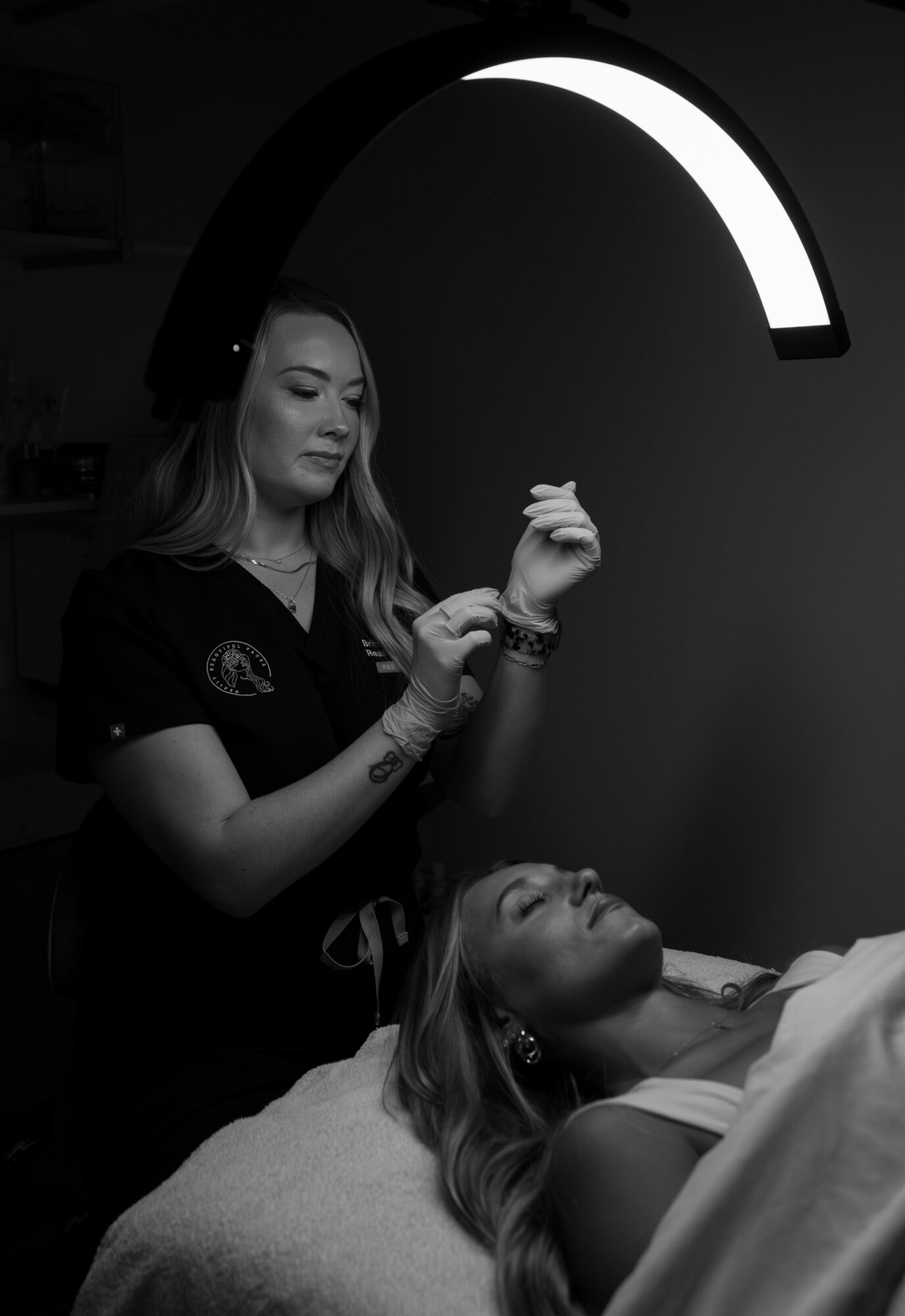 A woman in a dark uniform stands beside a reclining woman in a medical setting, under a curved light fixture.