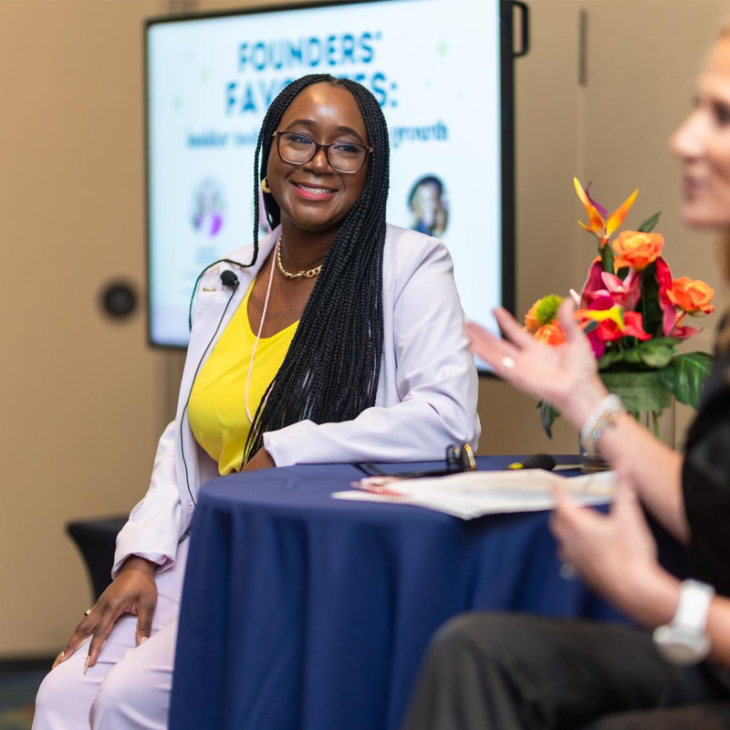 Smiling woman with glasses and long braids in a white blazer and yellow top, sitting at a table with flowers, during a discussion.