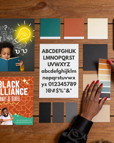 Person's hand with red nails reaching for a color palette on a wooden table, surrounded by color samples, alphabet cards, and a book.