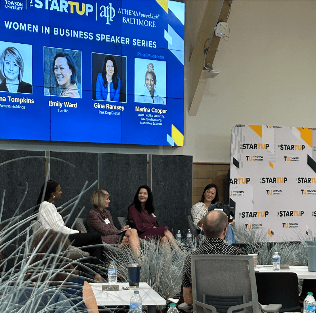 Panel discussion at Women in Business Speaker Series with four women seated on stage, large screen displaying speaker names, and audience members listening.