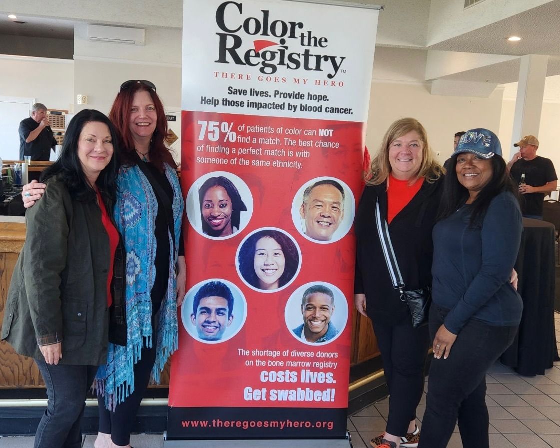Four women standing next to a banner promoting blood cancer awareness, smiling indoors.