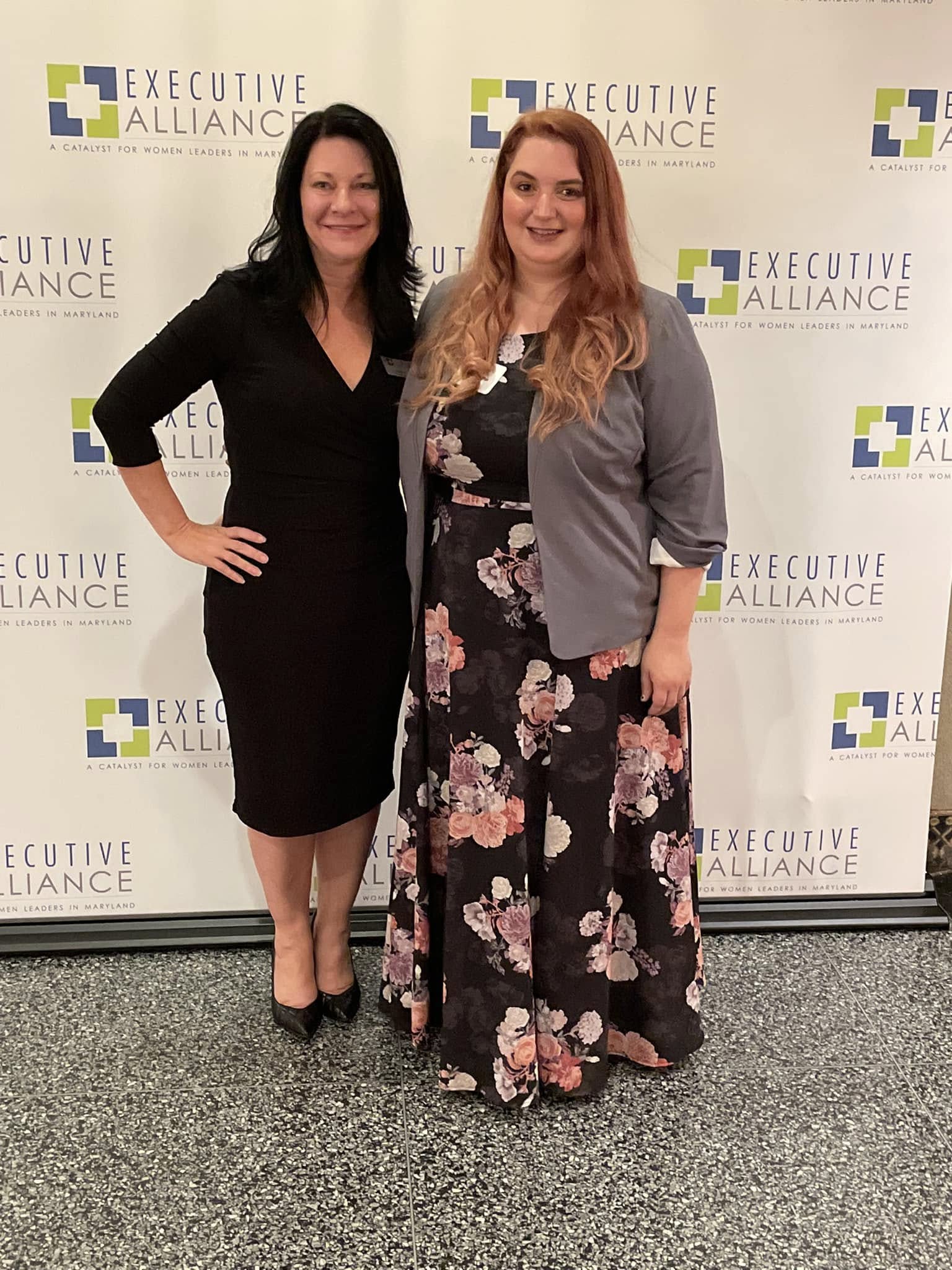 Two women standing side by side in front of a backdrop with 'Executive Alliance' logo, smiling, one in a black dress, the other in a floral dress and gray blazer.