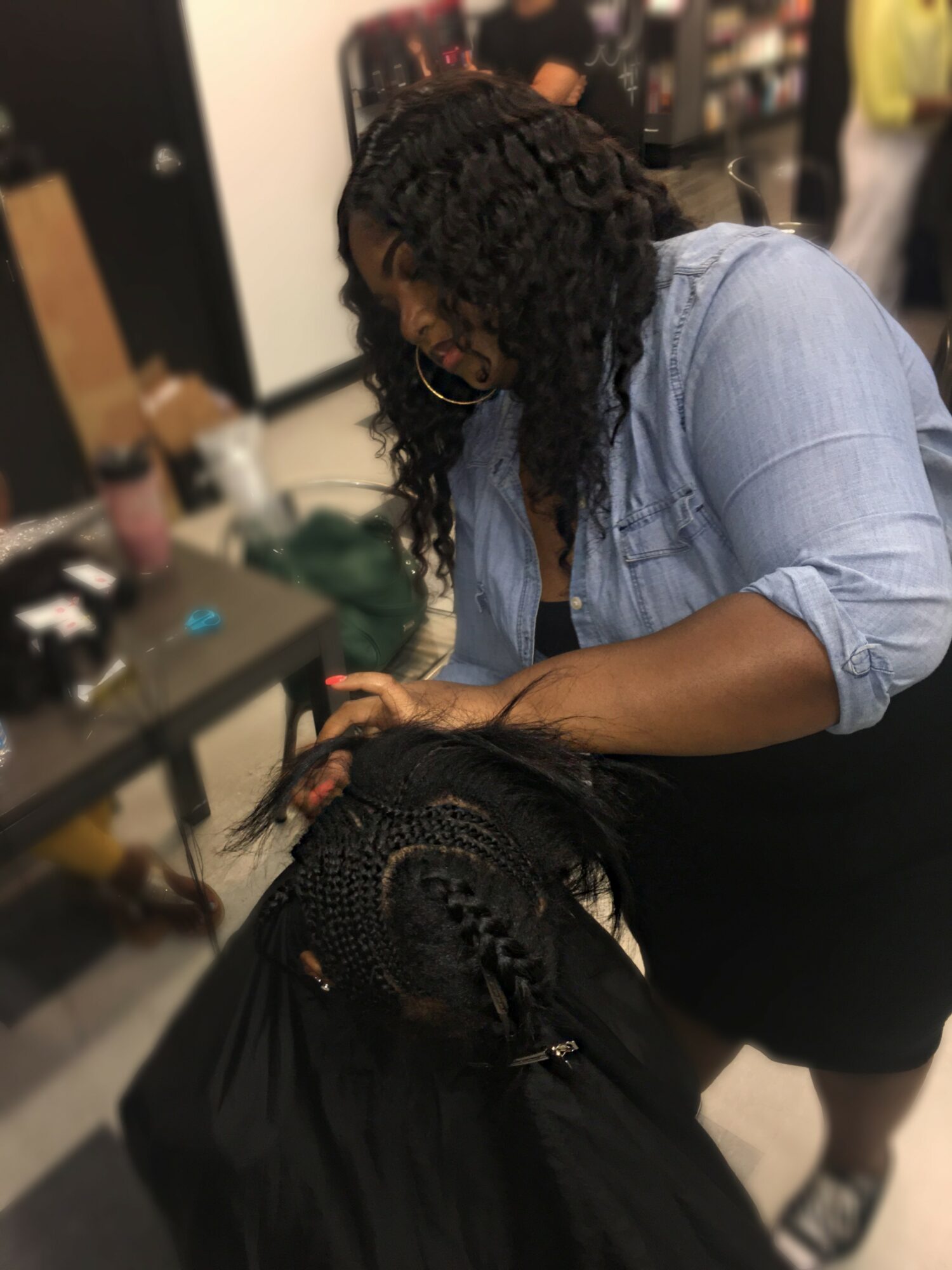 Woman with curly hair styling a person's braided hair in a salon, with hair tools and products nearby.