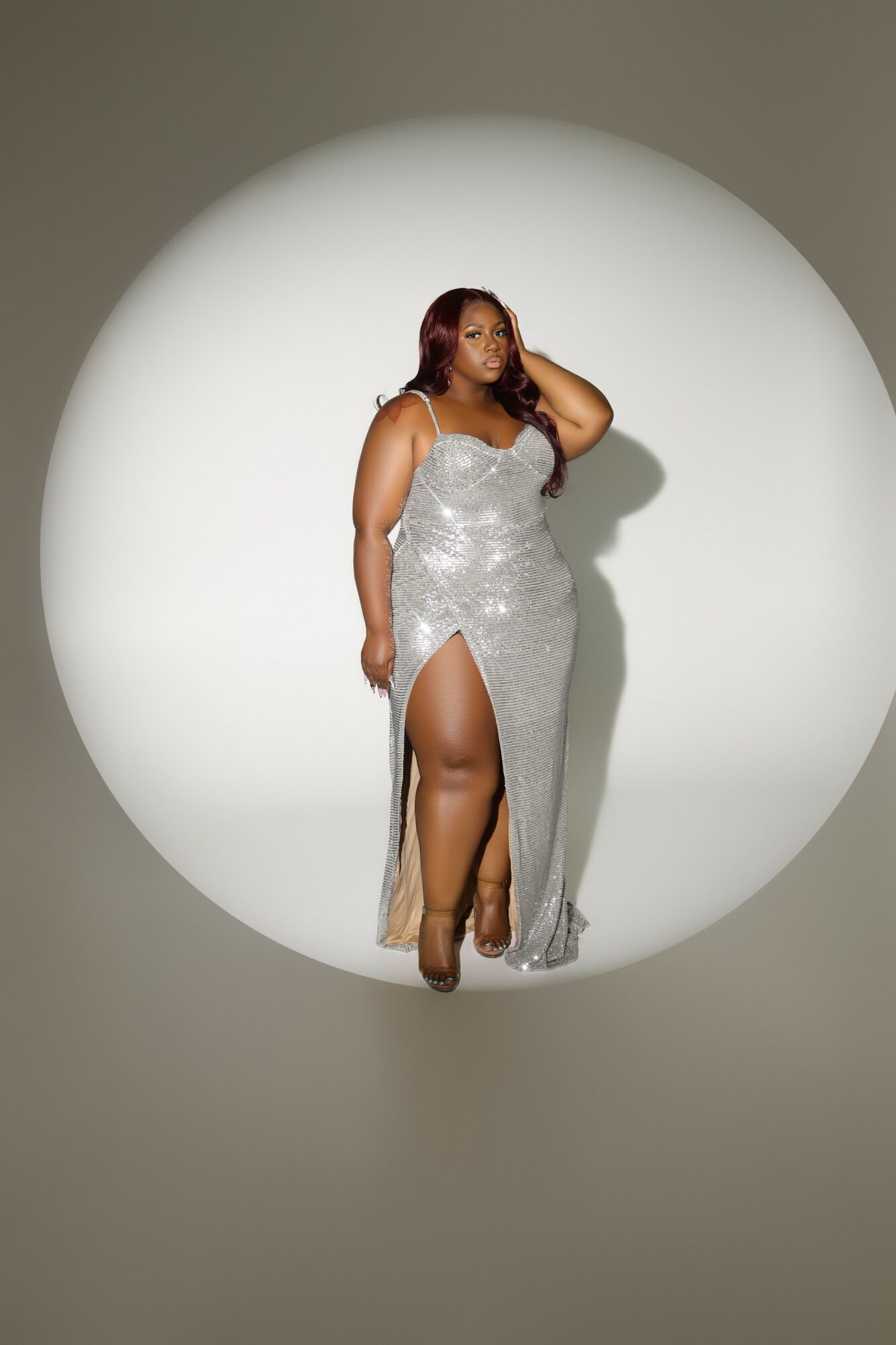 Woman in a silver, sequined gown with a high slit, standing in a spotlight against a plain background.