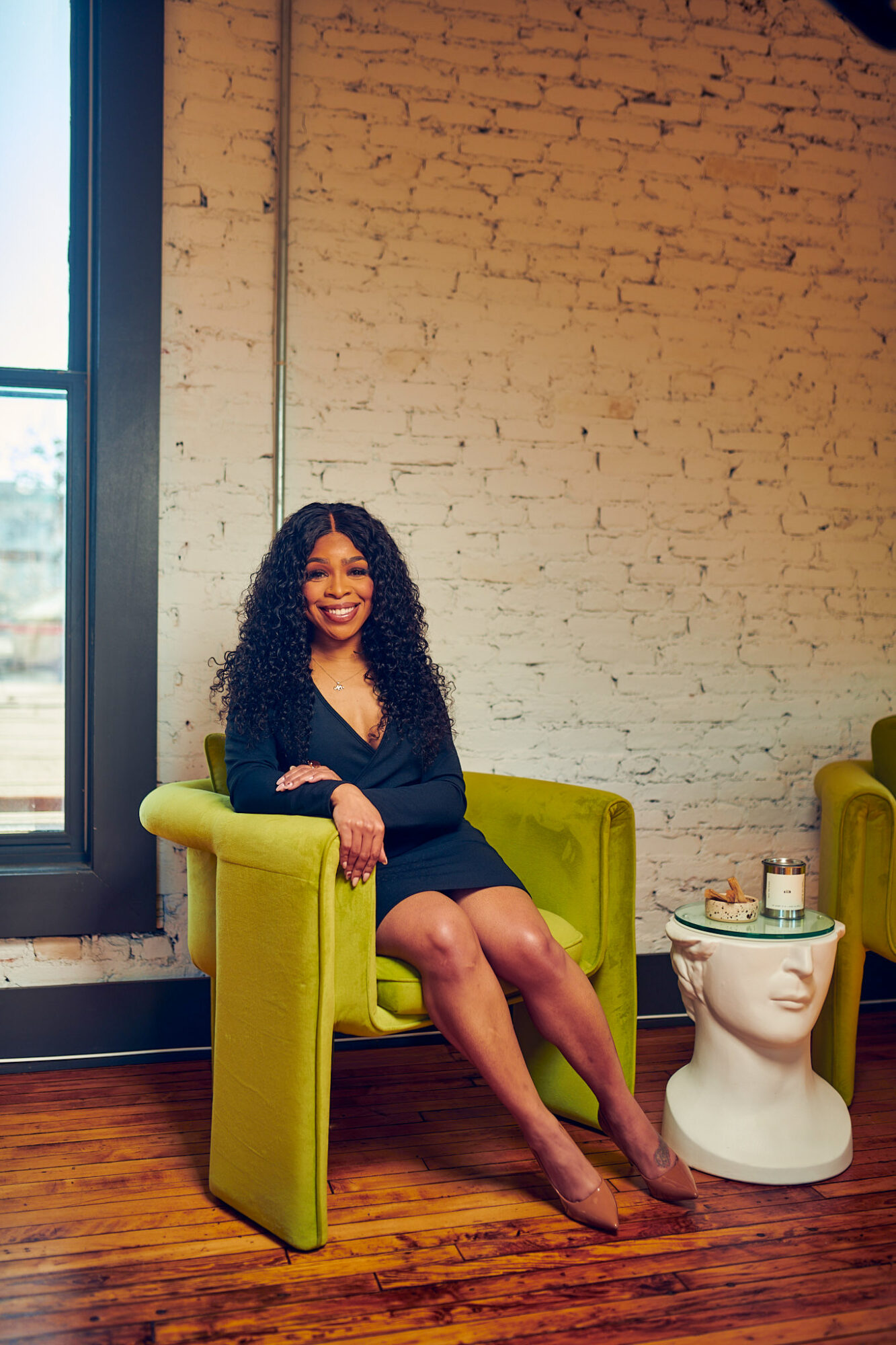 Woman with curly hair sitting on a green armchair in a room with a brick wall and wooden floor.