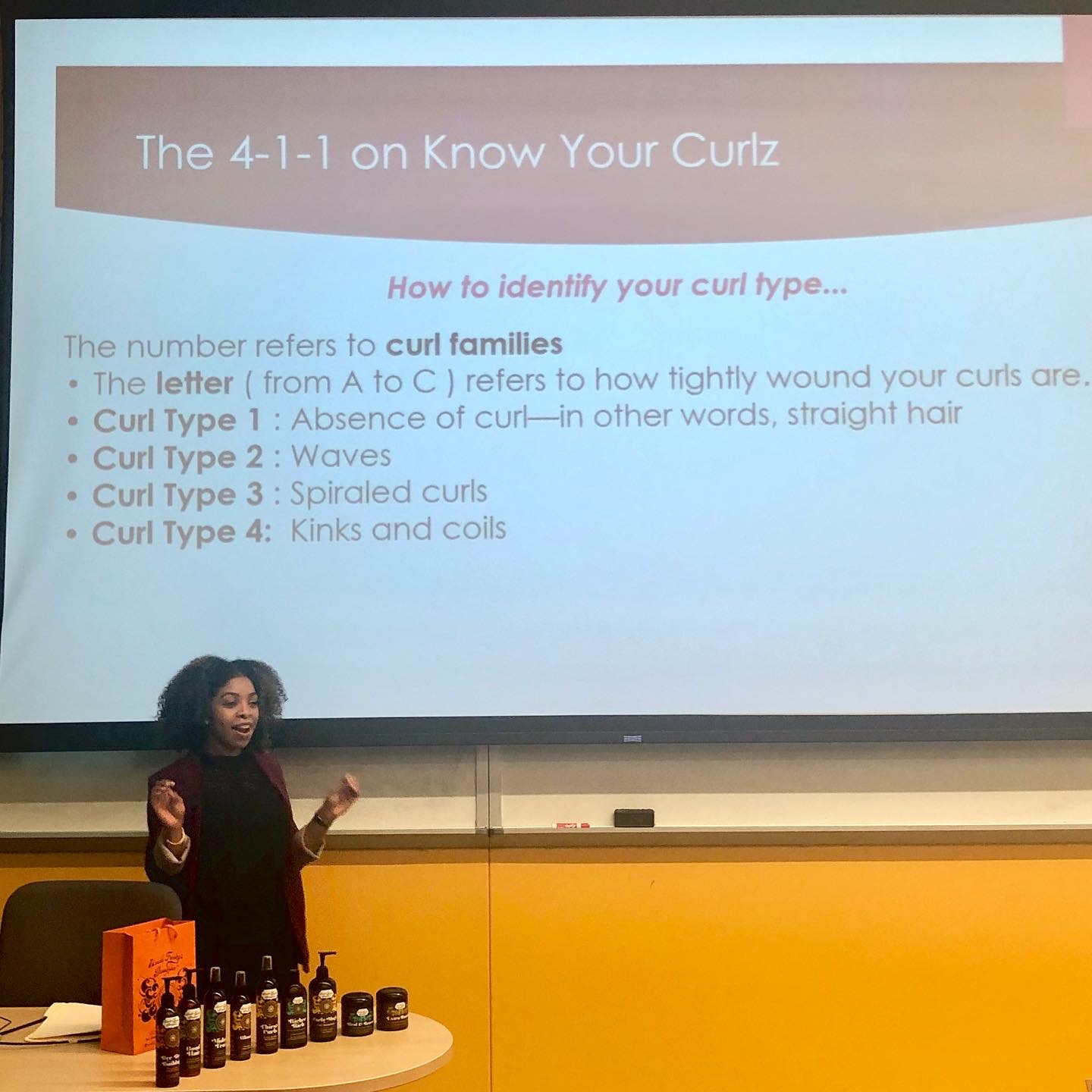 A woman stands in front of a presentation slide titled 'The 4-1-1 on Know Your Curlz,' explaining curl types with a pointer. The slide lists four curl types and their descriptions.
