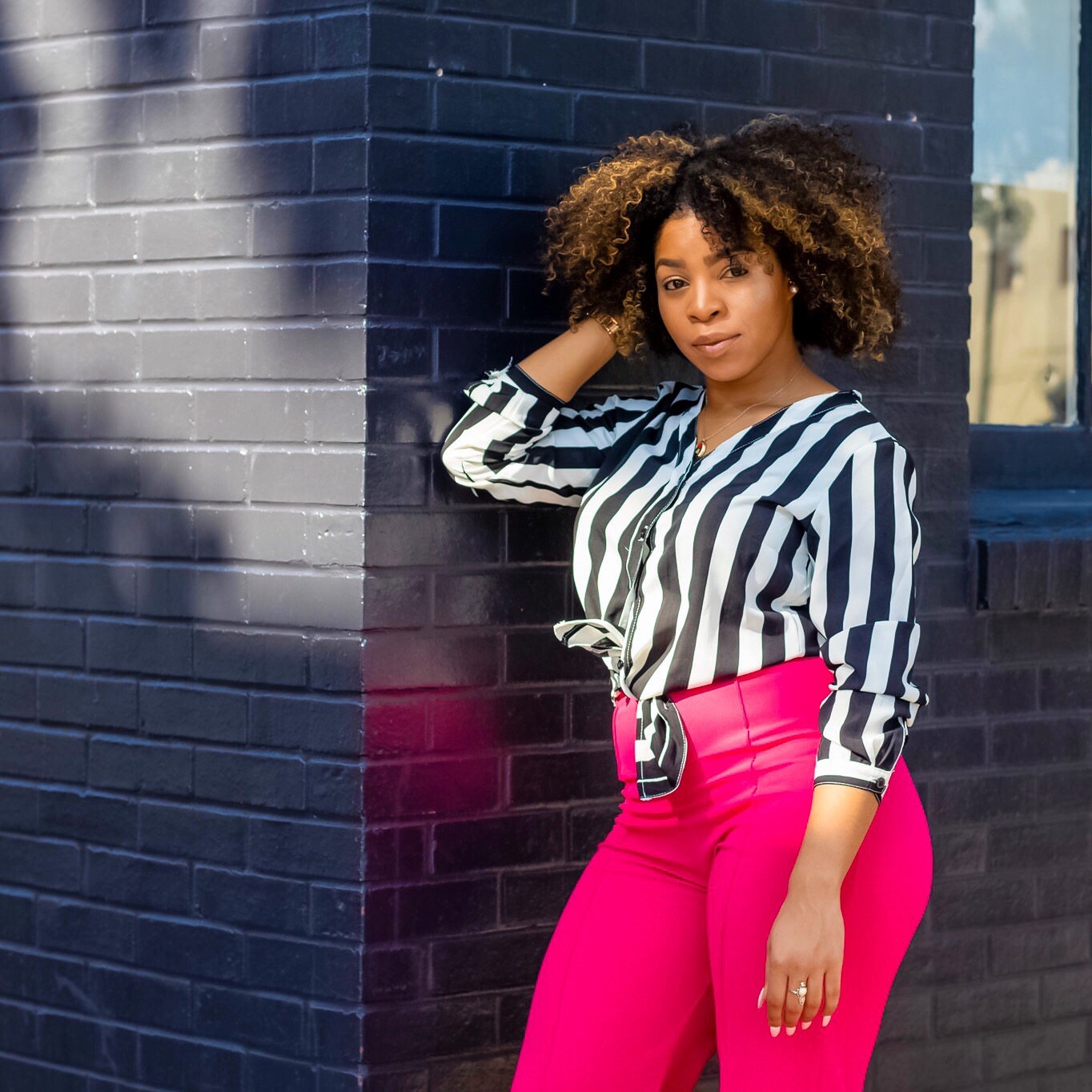 Woman with curly hair leaning against a dark brick wall, wearing a striped shirt and bright pink pants.