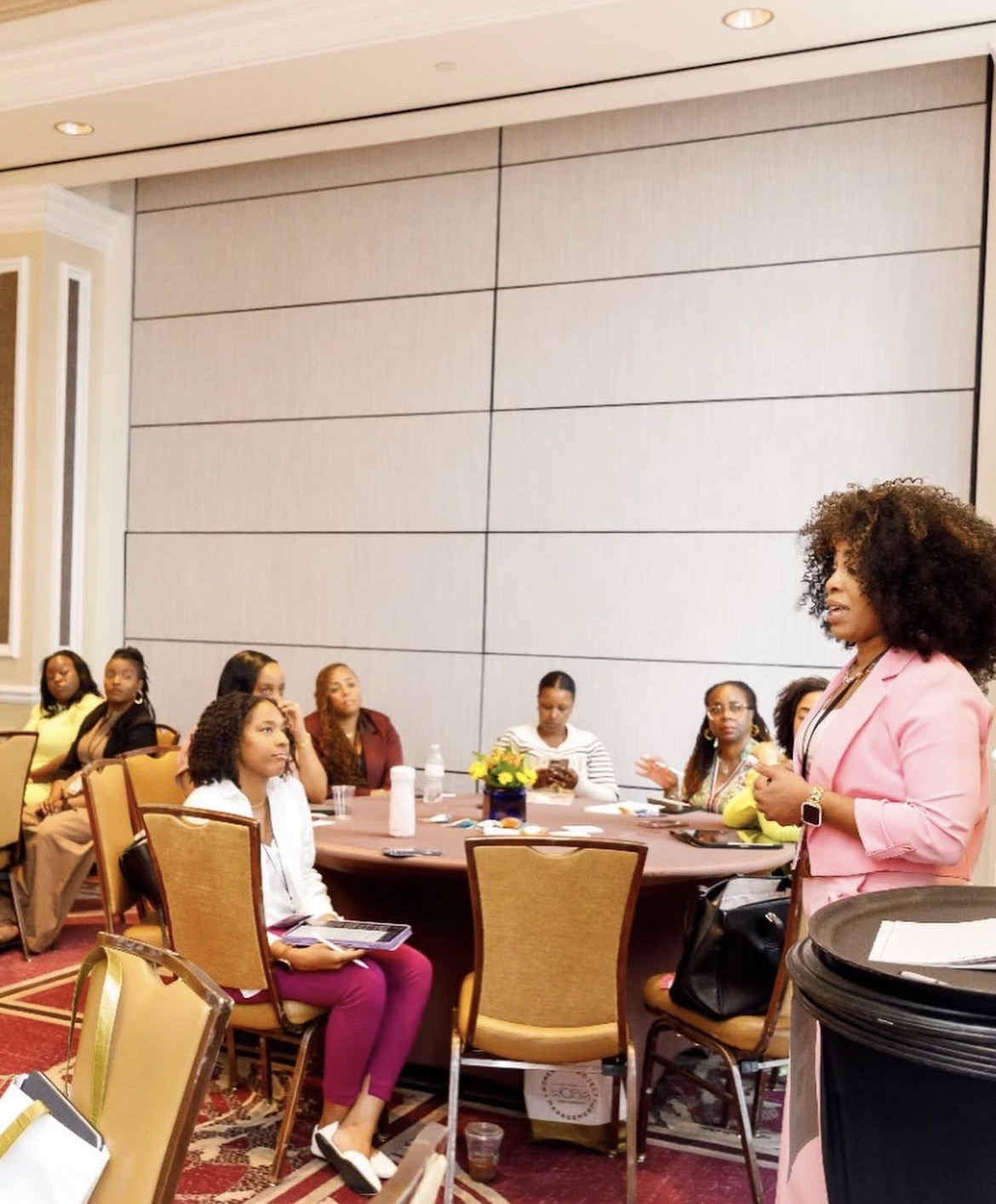 A woman in pink speaking to a group of children seated around a table in a conference room.