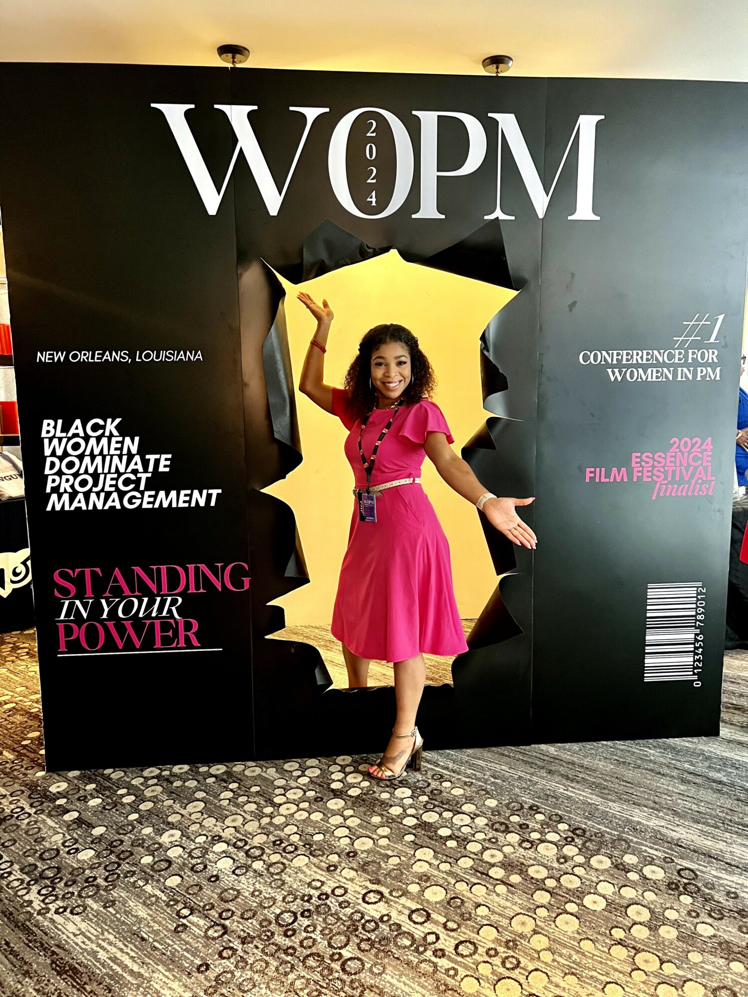 Woman in pink dress standing in front of a black backdrop with event text, holding a yellow star-shaped prop, smiling.
