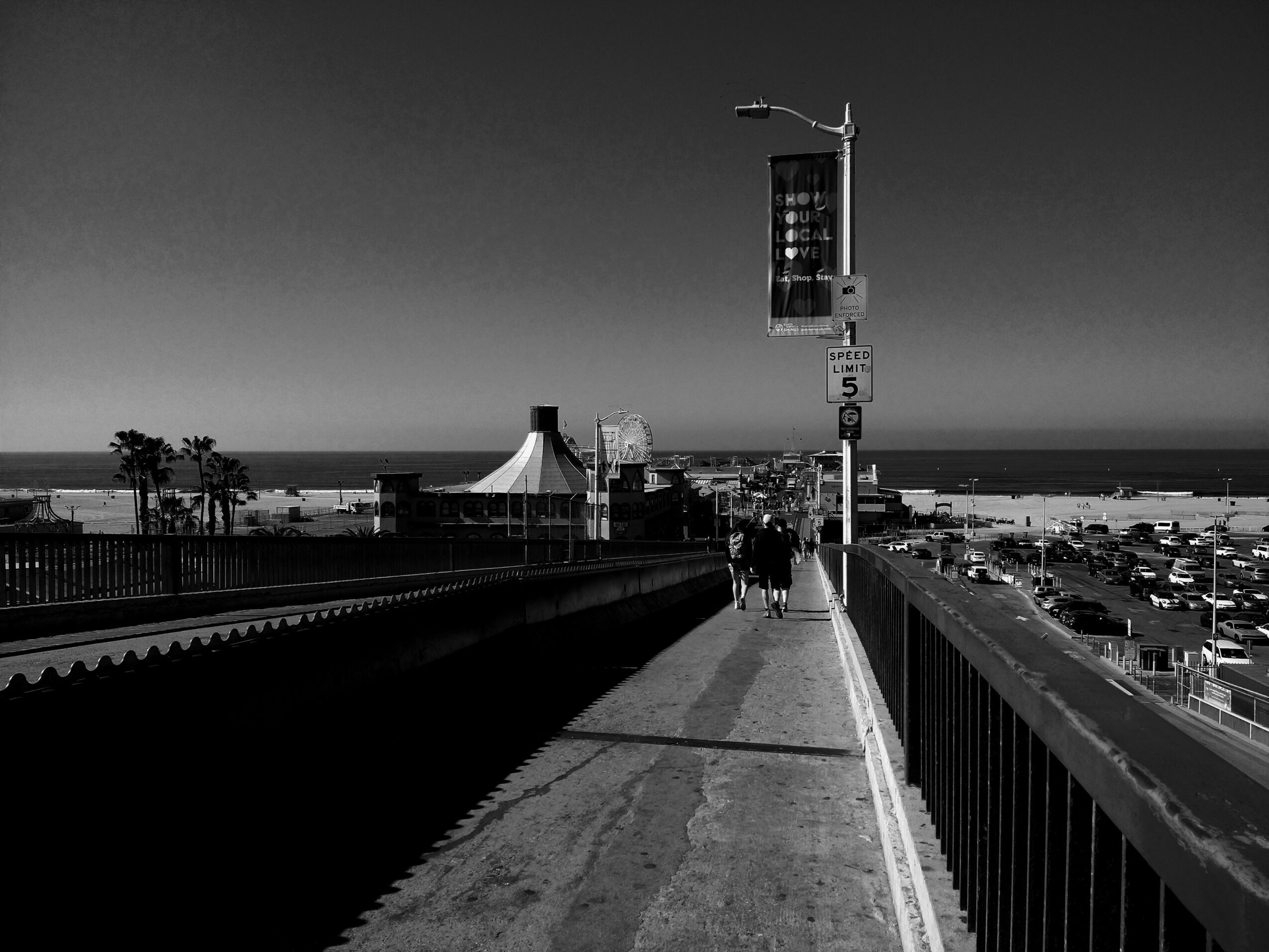 People walk along a pier with a tent structure, a lamppost, and a parking lot in the background, under a clear sky.