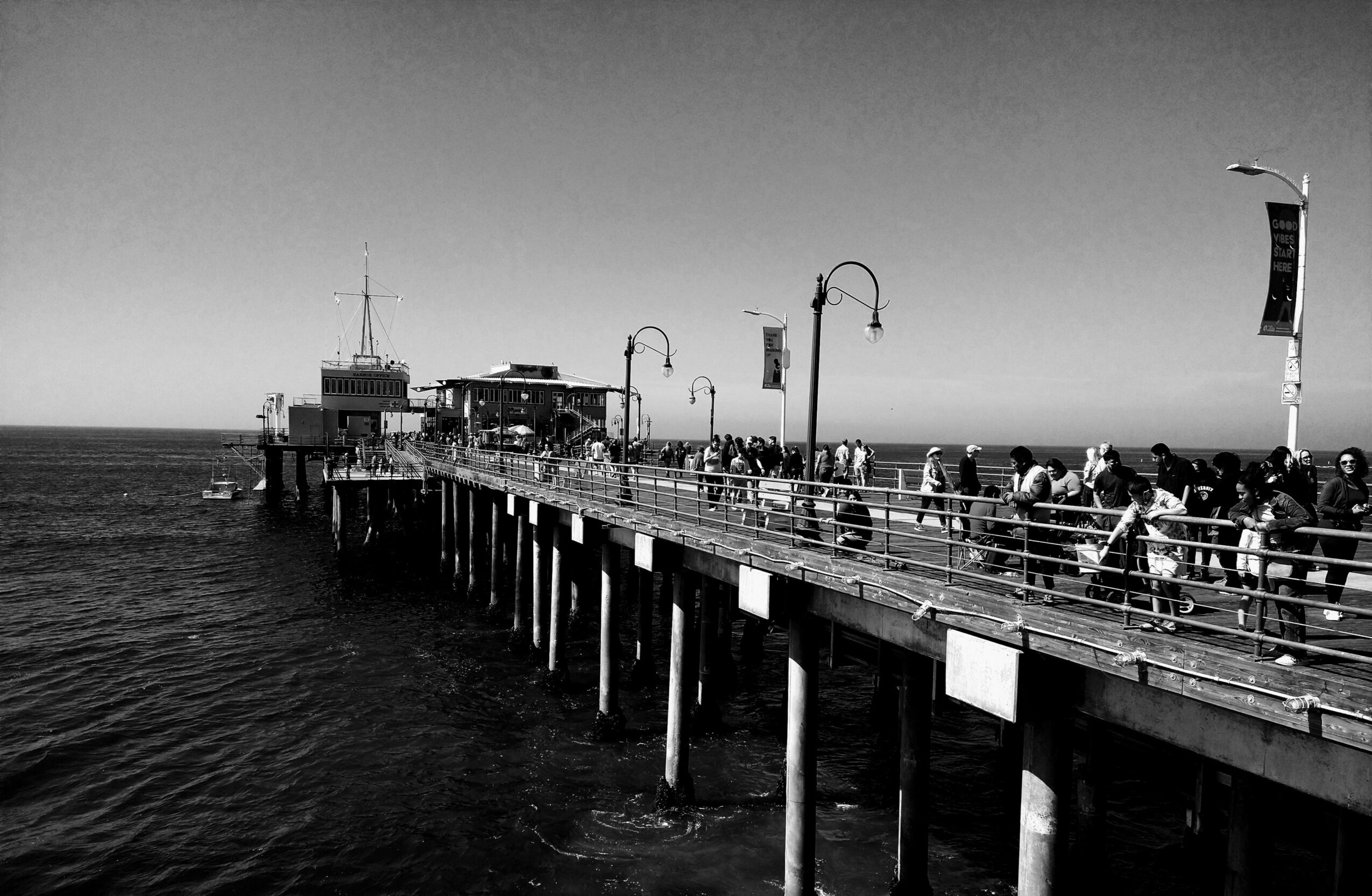 Pier extending into the water with people walking, lampposts, and a small building at the end, in black and white.