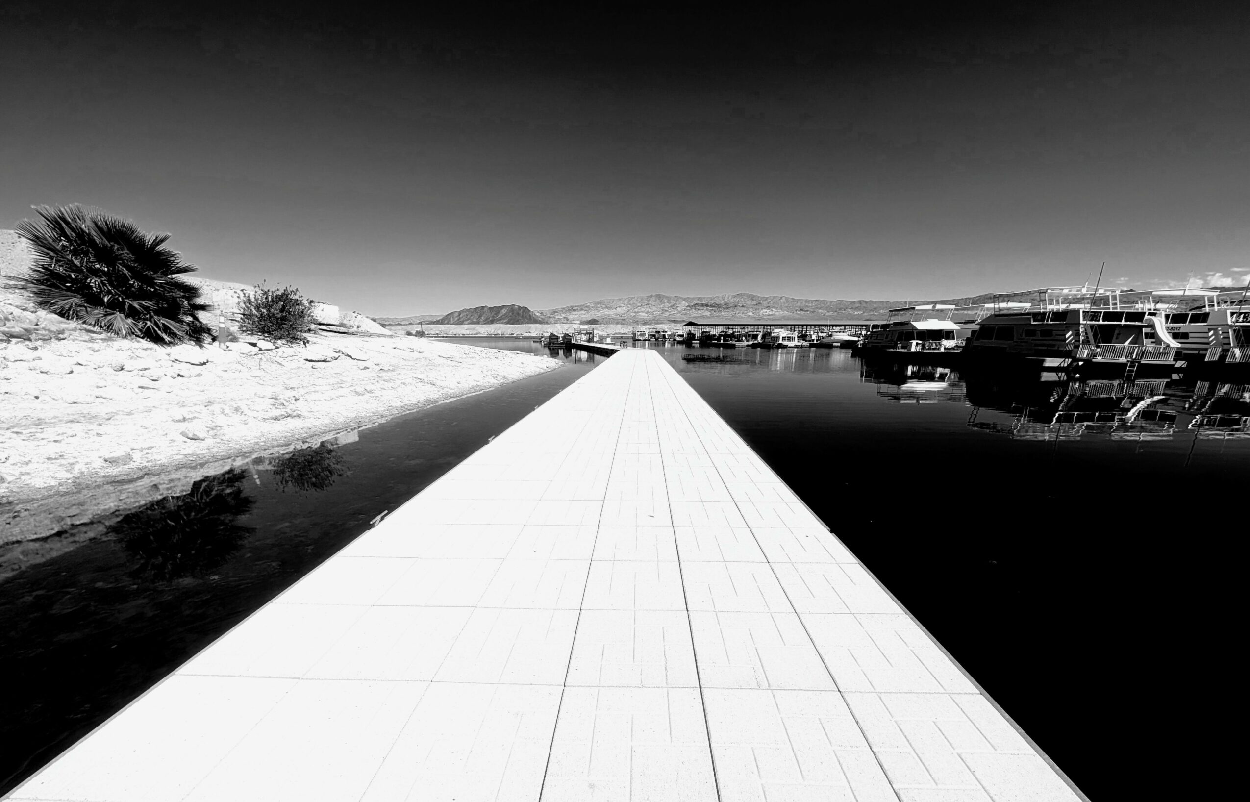 Pathway leading to a harbor with boats, snow-covered landscape on the left, water on the right, and a clear sky.