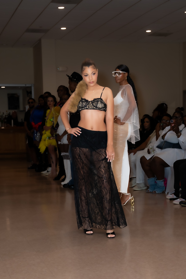 Young woman walking on runway in black lace skirt and strapless top, audience seated on sides, indoor event