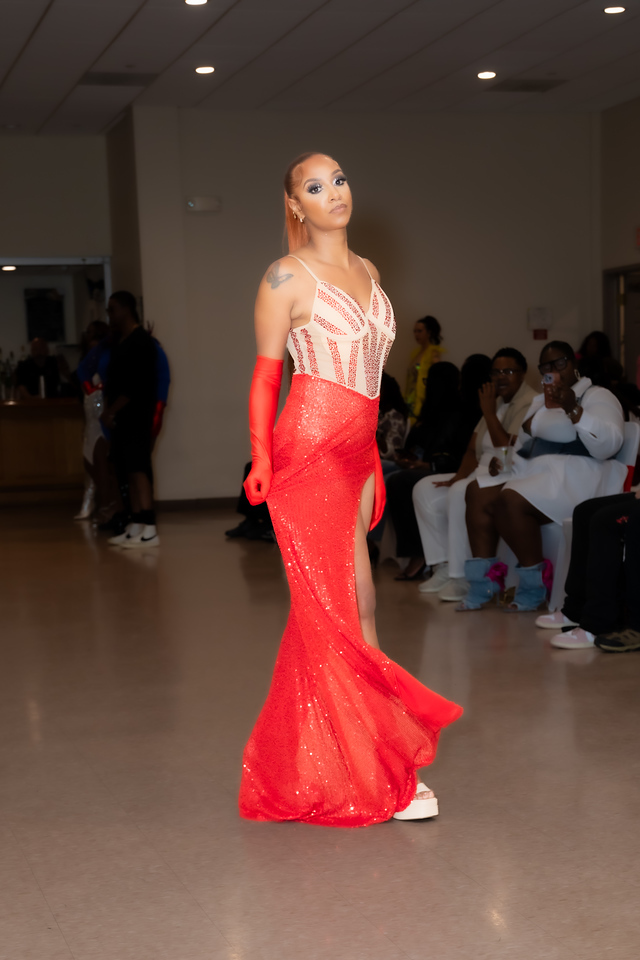 Woman in a red gown with sheer top and long gloves, standing in a room with seated people in the background.