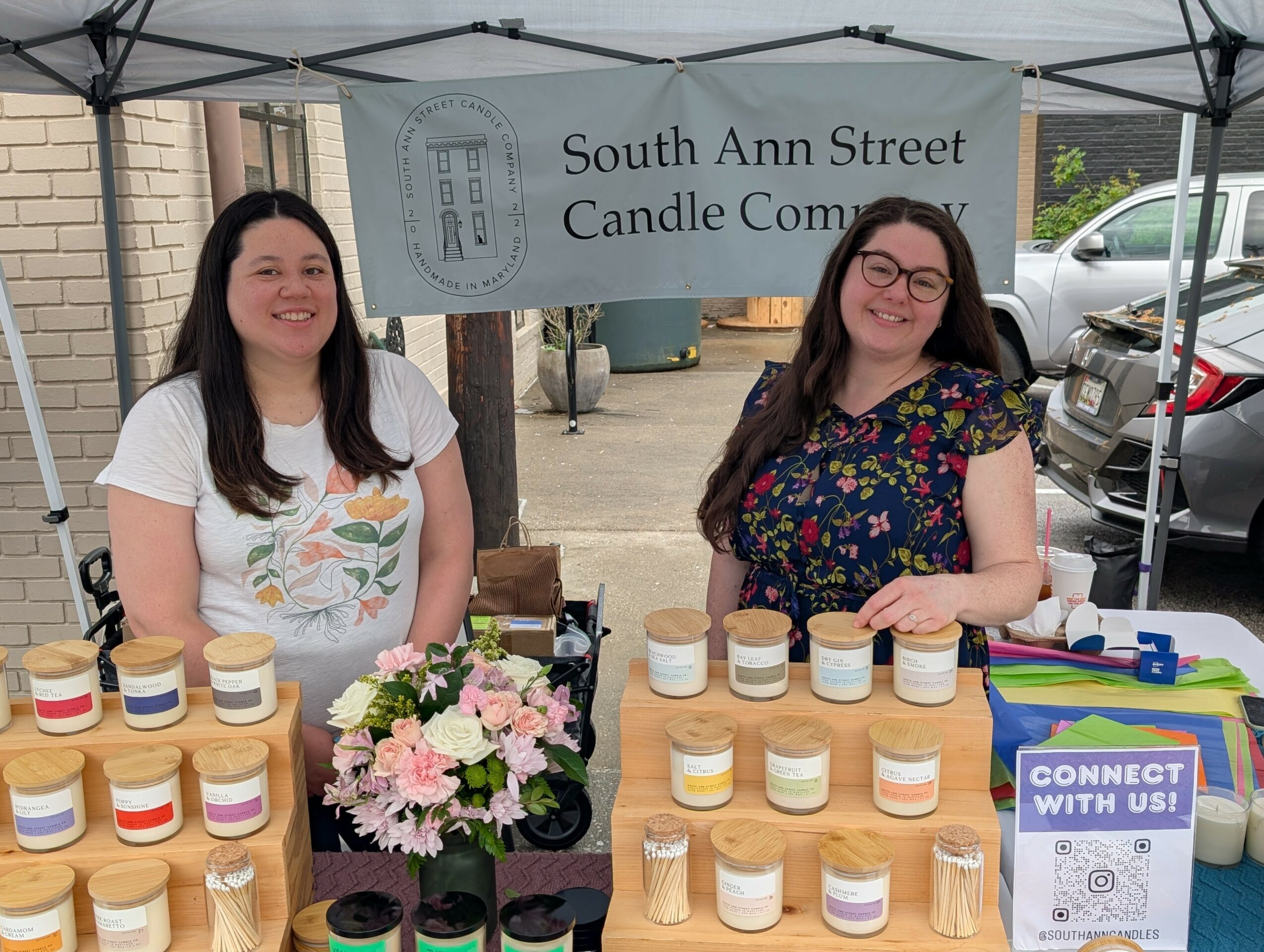 Two women stand behind a table with candles, flowers, and a sign for South Ann Street Candle Company, outdoors.