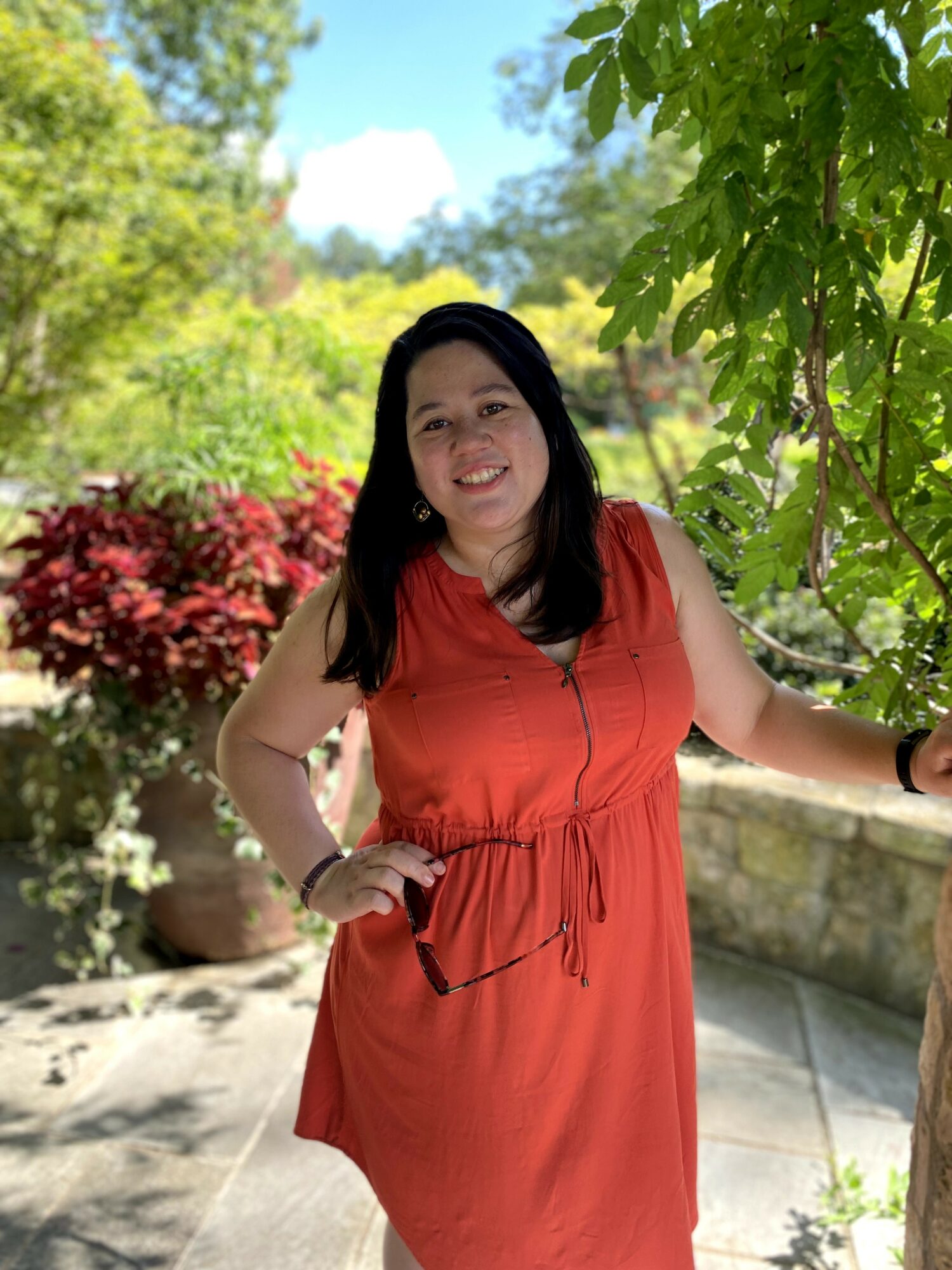 Woman in red dress standing outdoors with greenery and trees, smiling, holding a branch, sunny day.