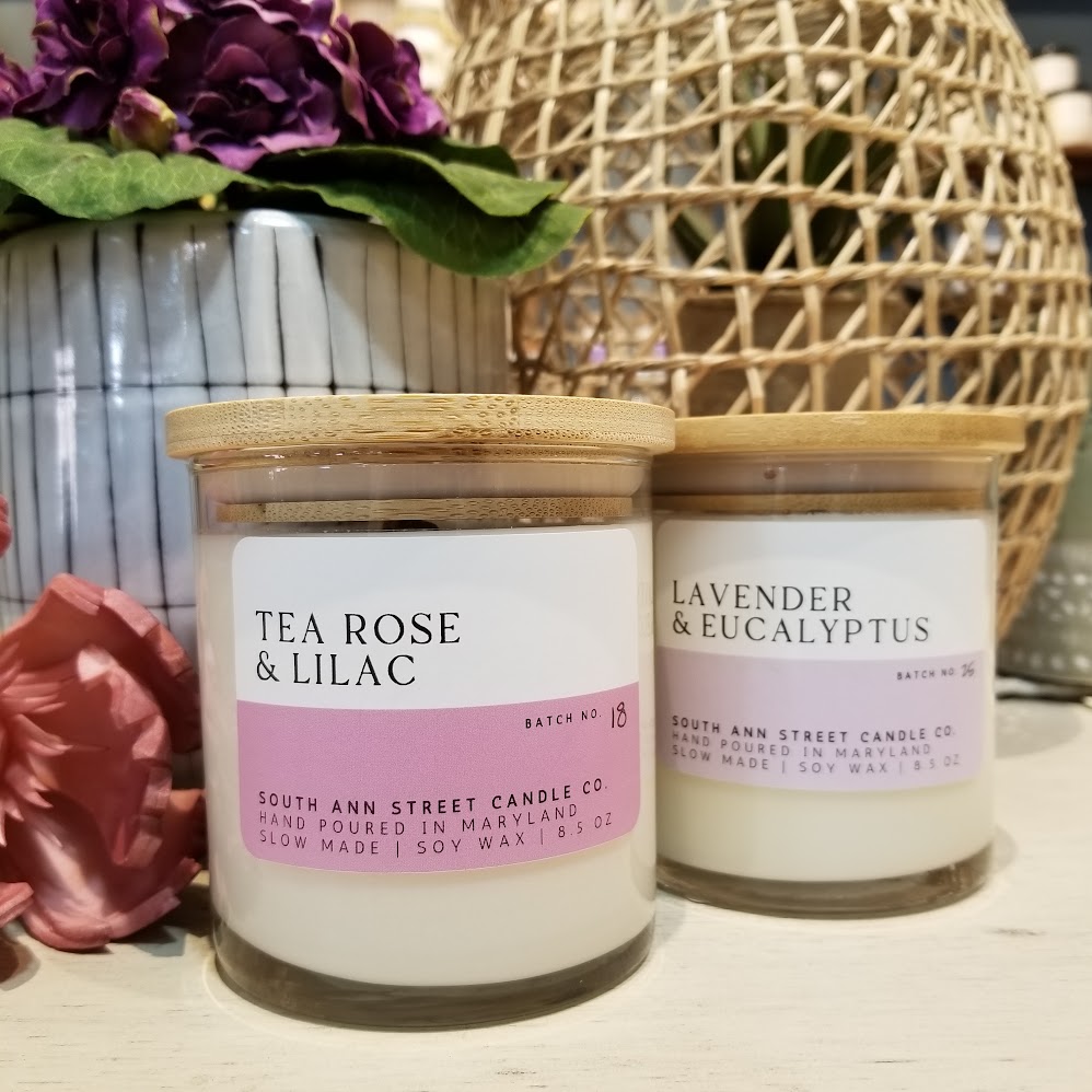 Two candles with wooden lids, one labeled lavender and eucalyptus, the other tea rose and lilac, on a table with decorative items.