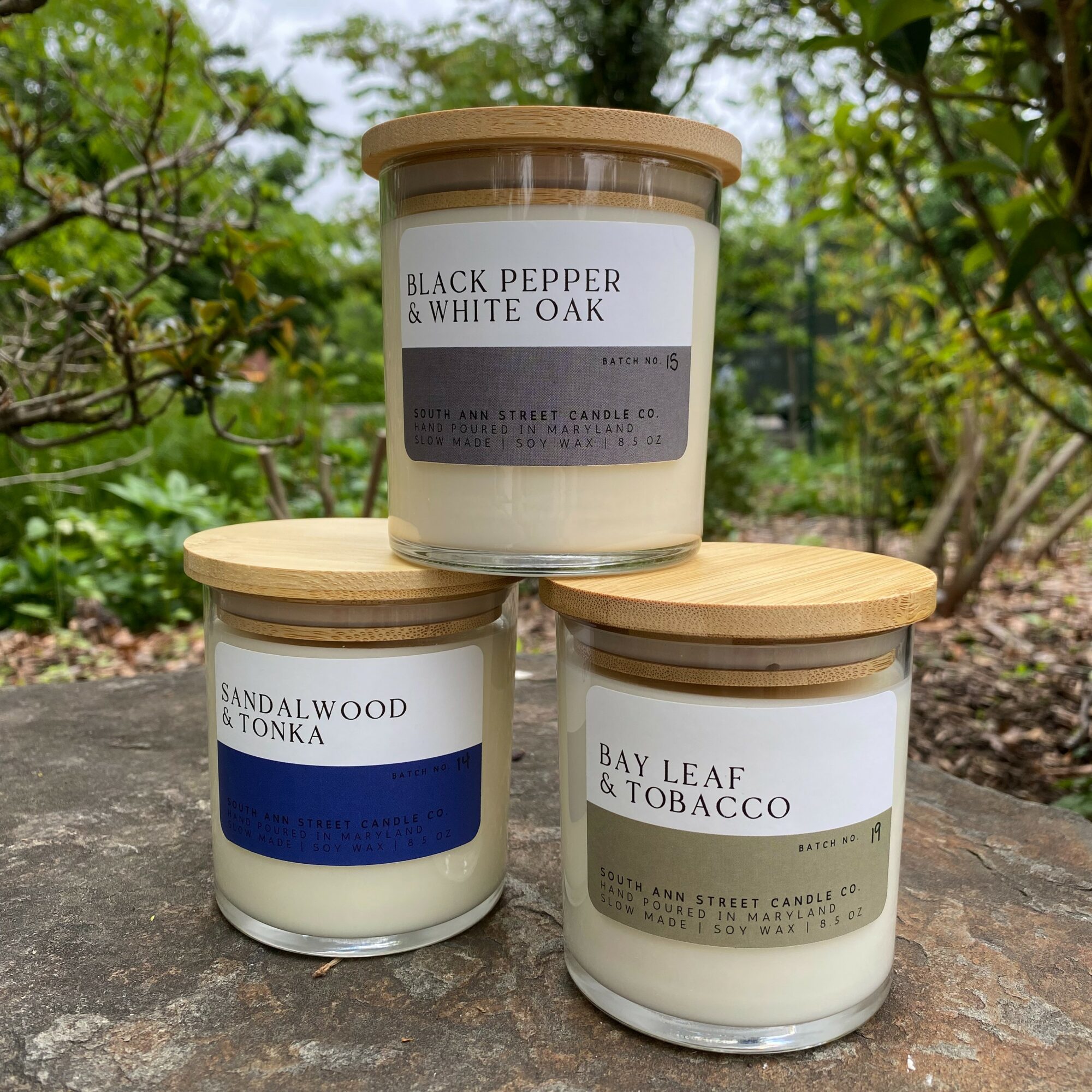 Three candles outdoors on a rock, with labels reading black pepper, sandalwood, and bay leaf, surrounded by greenery.