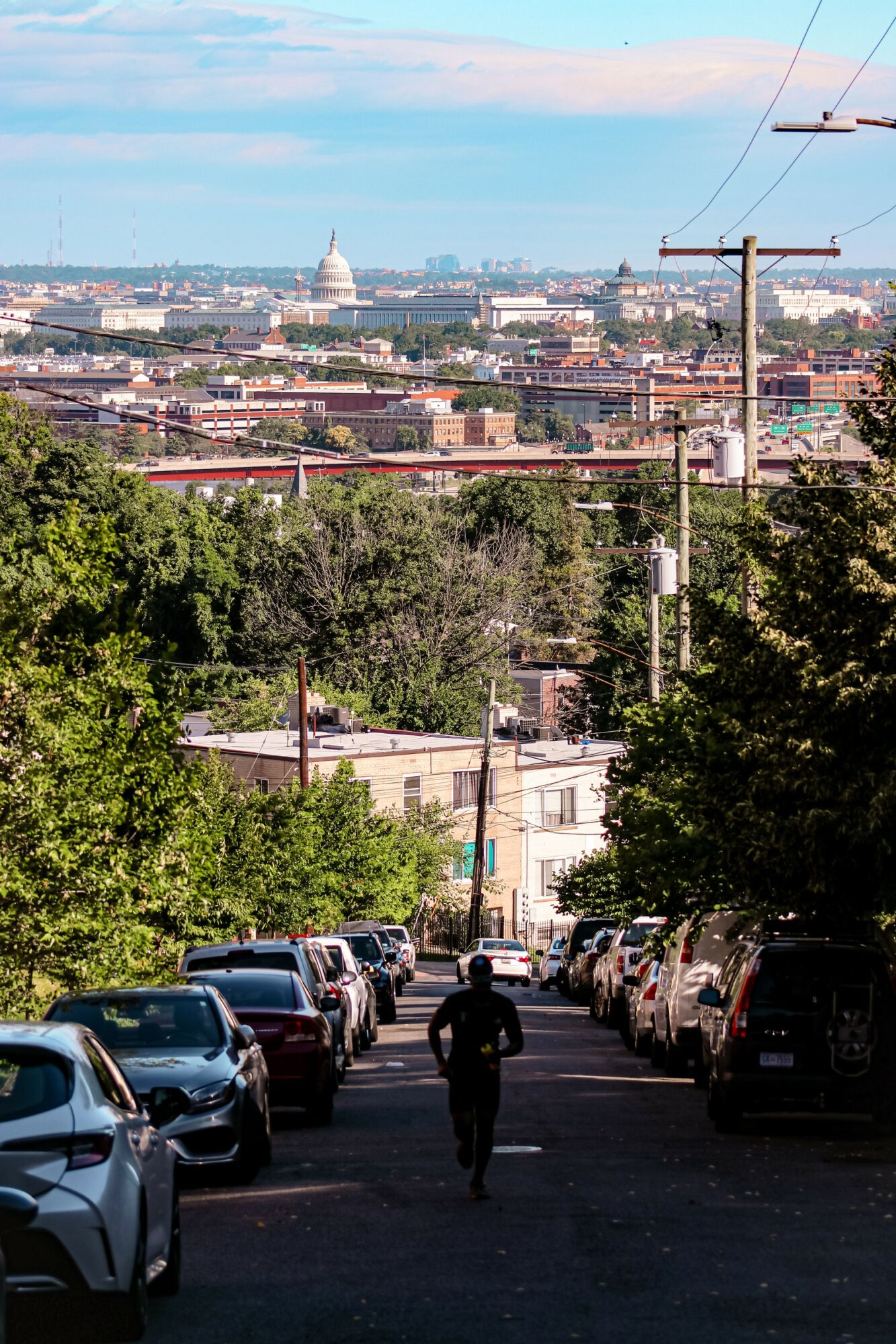 Person walking down a tree-lined street with parked cars, cityscape, and Capitol building in the background.