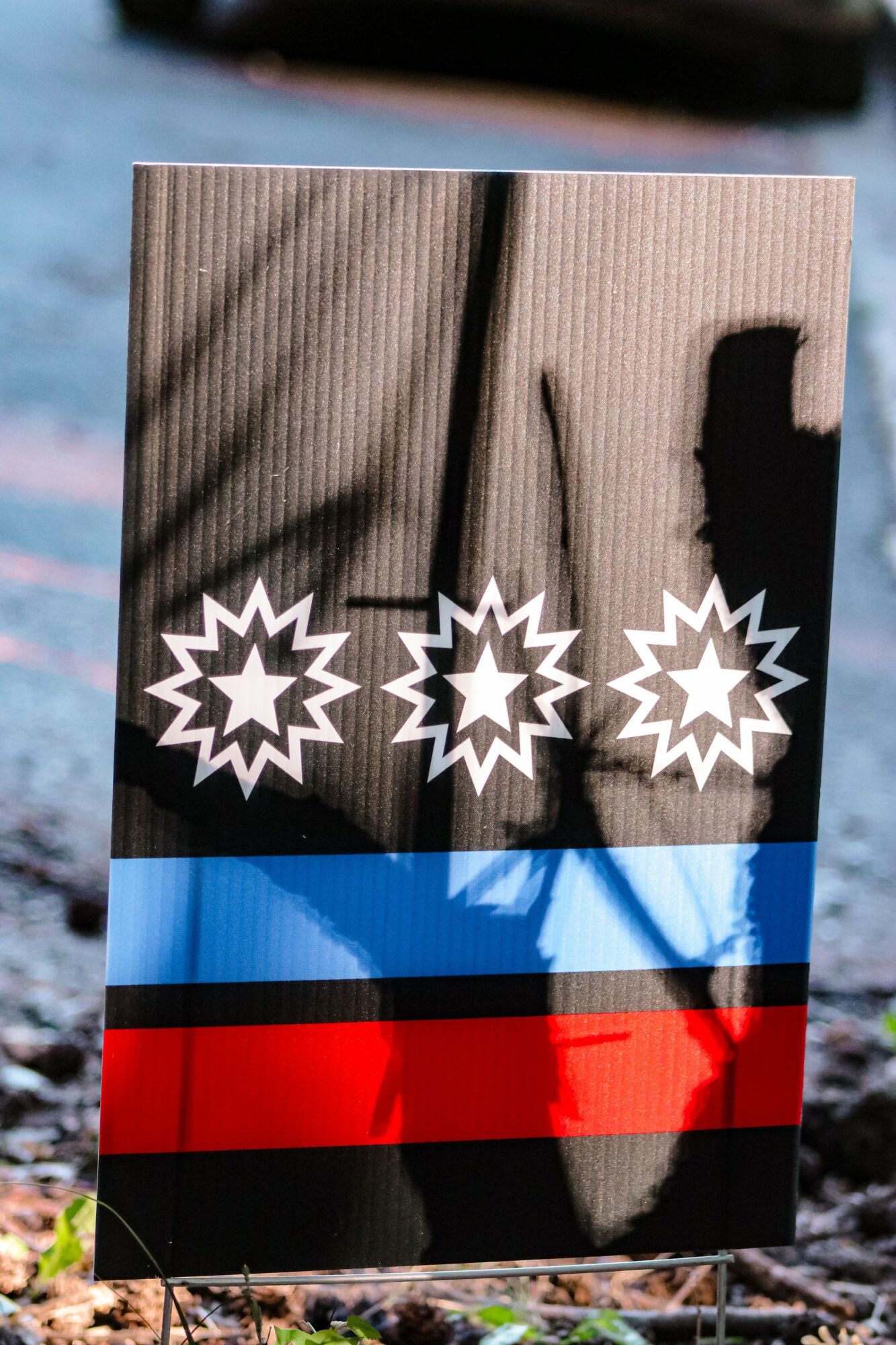 A wooden sign with three starburst symbols and horizontal blue and red stripes, outdoors with shadows and a blurred background.