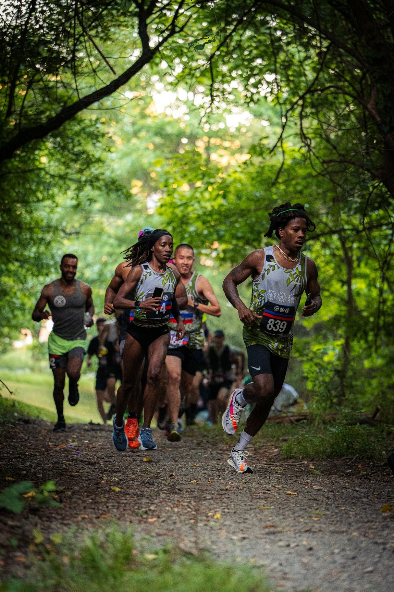 Group of runners participating in a race through a lush green forest, some wearing race bibs and athletic gear.