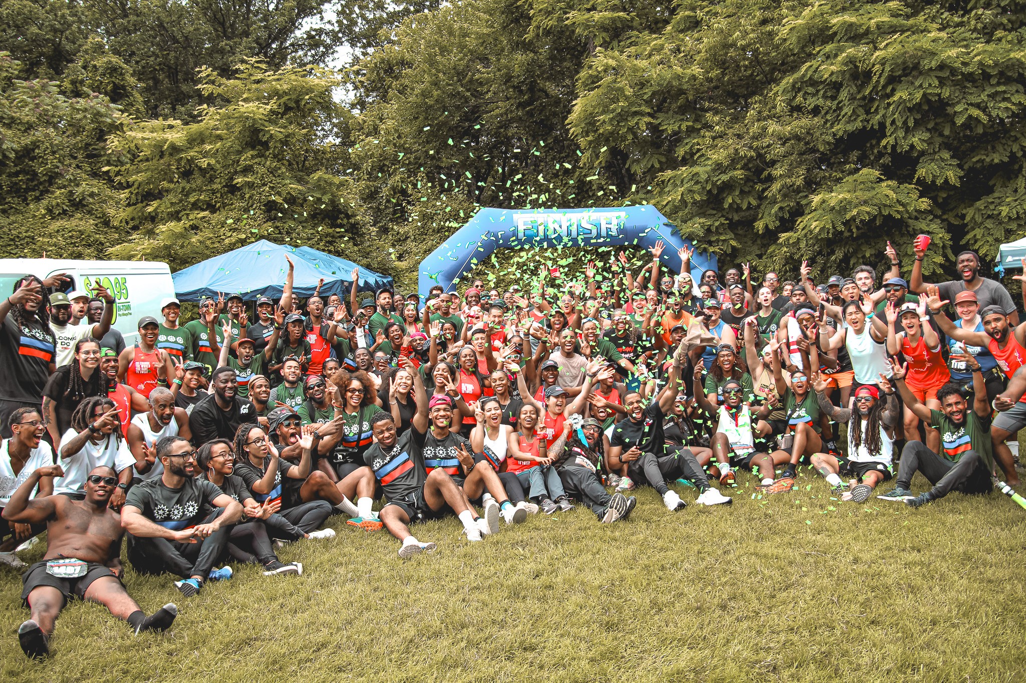 Large group of people outdoors on grass, celebrating with confetti, under a blue inflatable arch, trees in background.