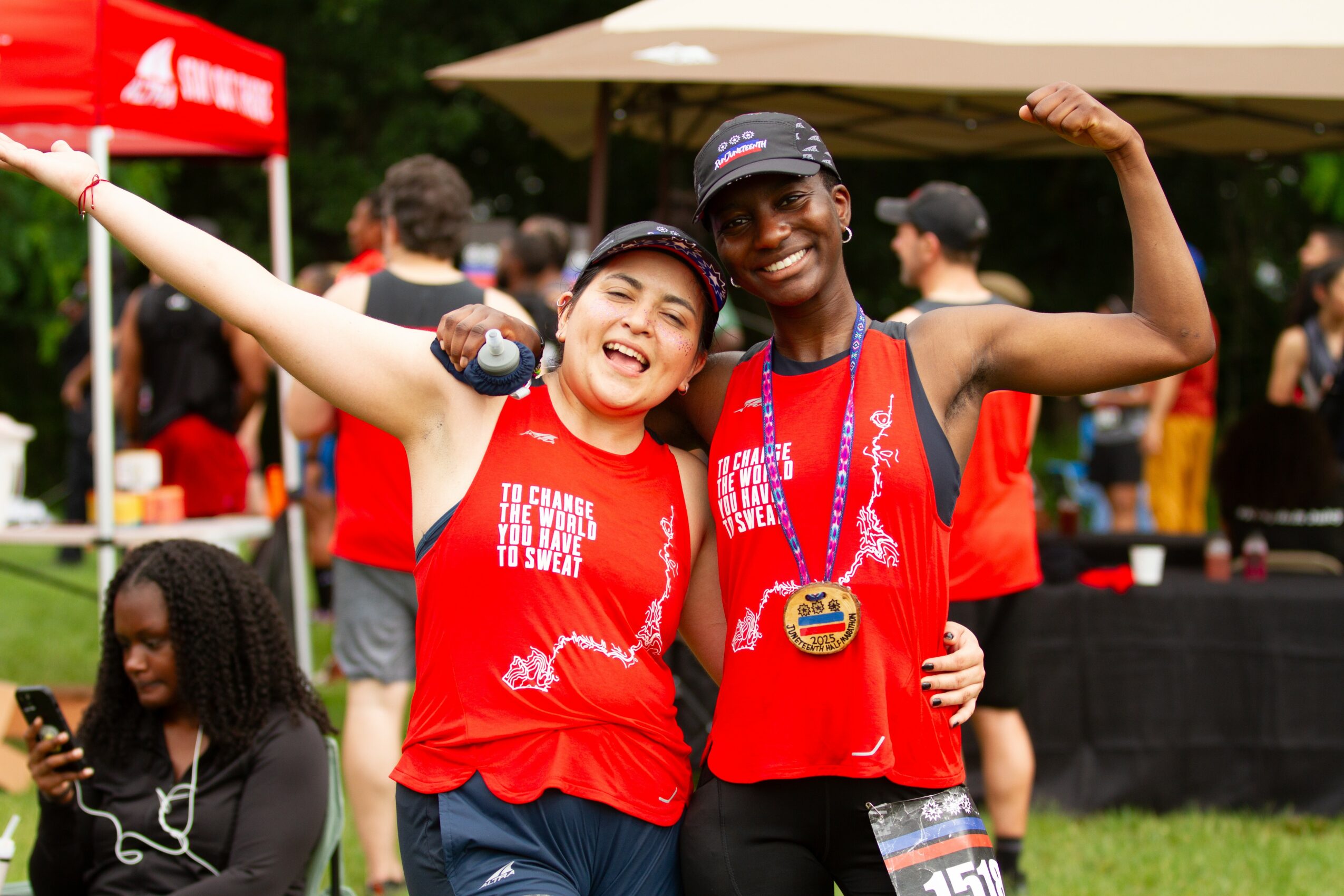 Two smiling women in red shirts, one with a medal, celebrating outdoors with arms raised, surrounded by people and tents.