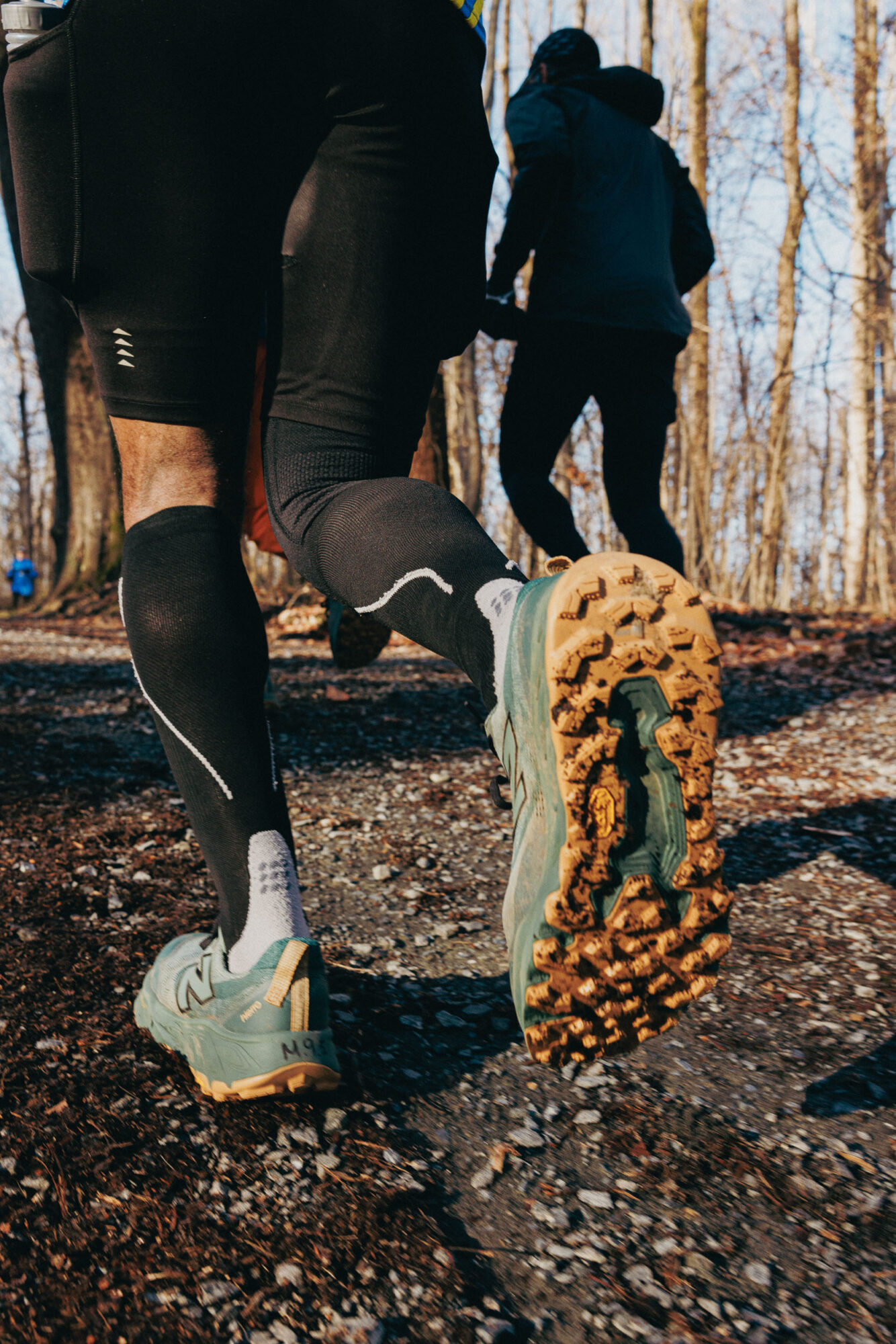 Close-up of a person running on a trail, focusing on their shoe and lower leg, with other runners in the background.