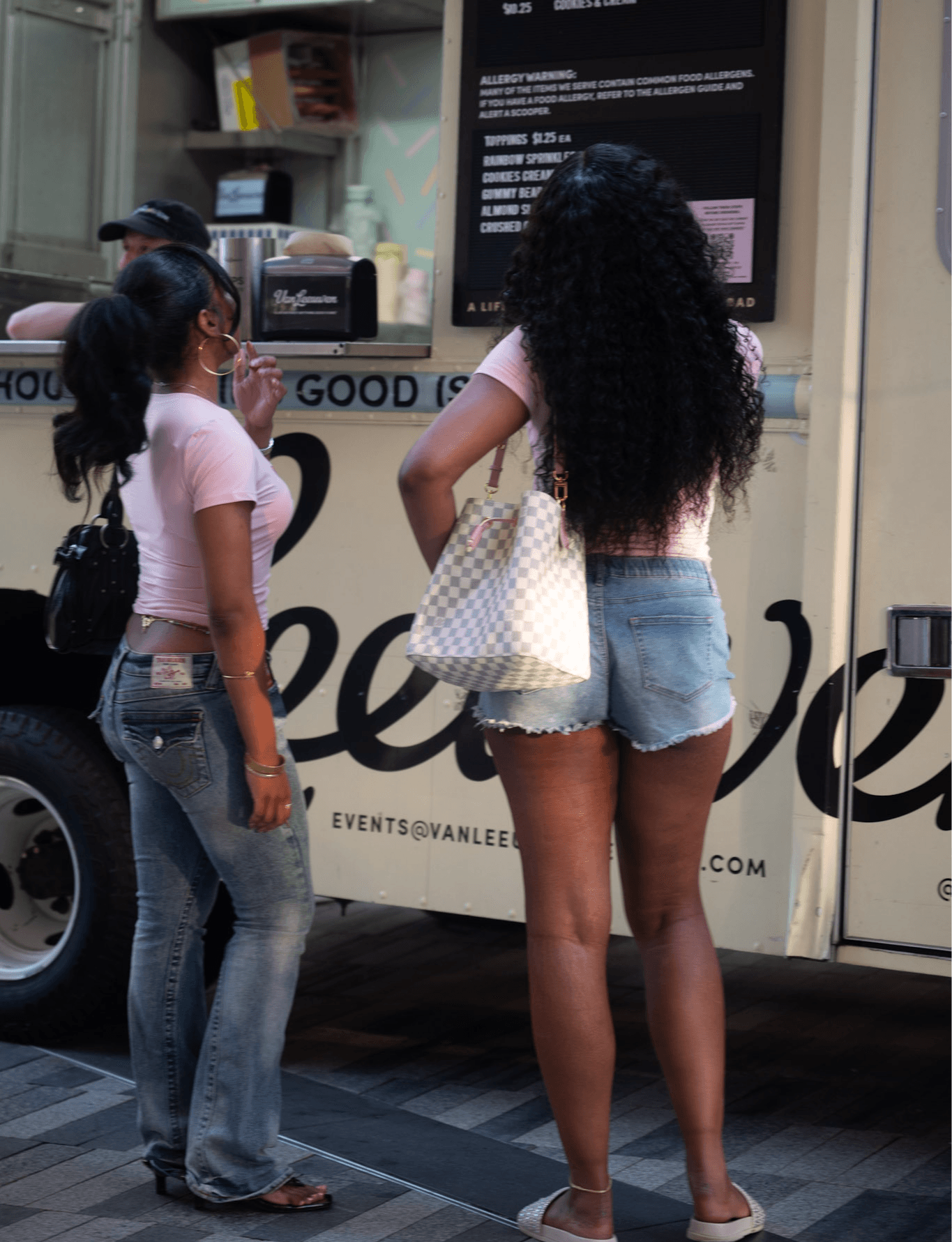 Two women with long curly hair stand near a food truck, one wearing a pink shirt and denim shorts, the other in a pink shirt and jeans, holding a bag.