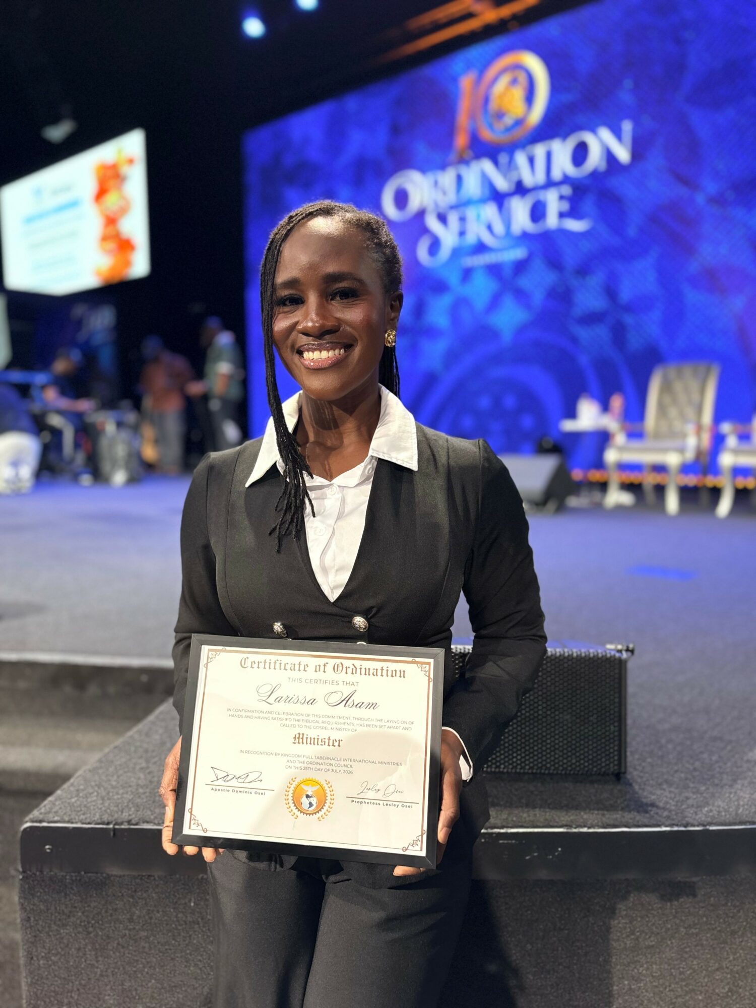 Smiling woman in formal attire holding a certificate on stage at an event, with a large screen displaying 'Observation Service' in the background.