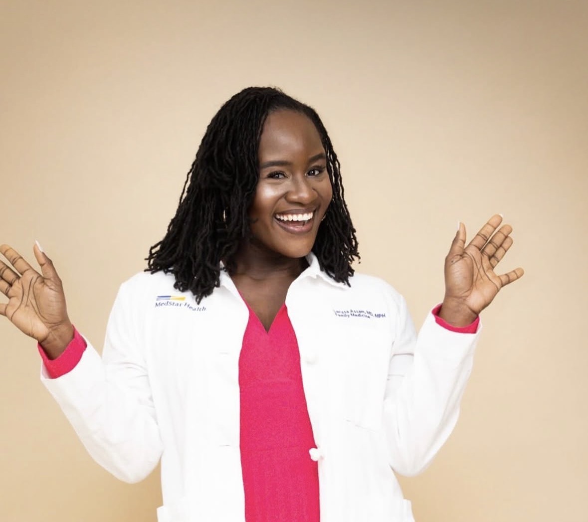 Smiling woman with shoulder-length dreadlocks wearing a white coat and pink shirt, raising hands, beige background.