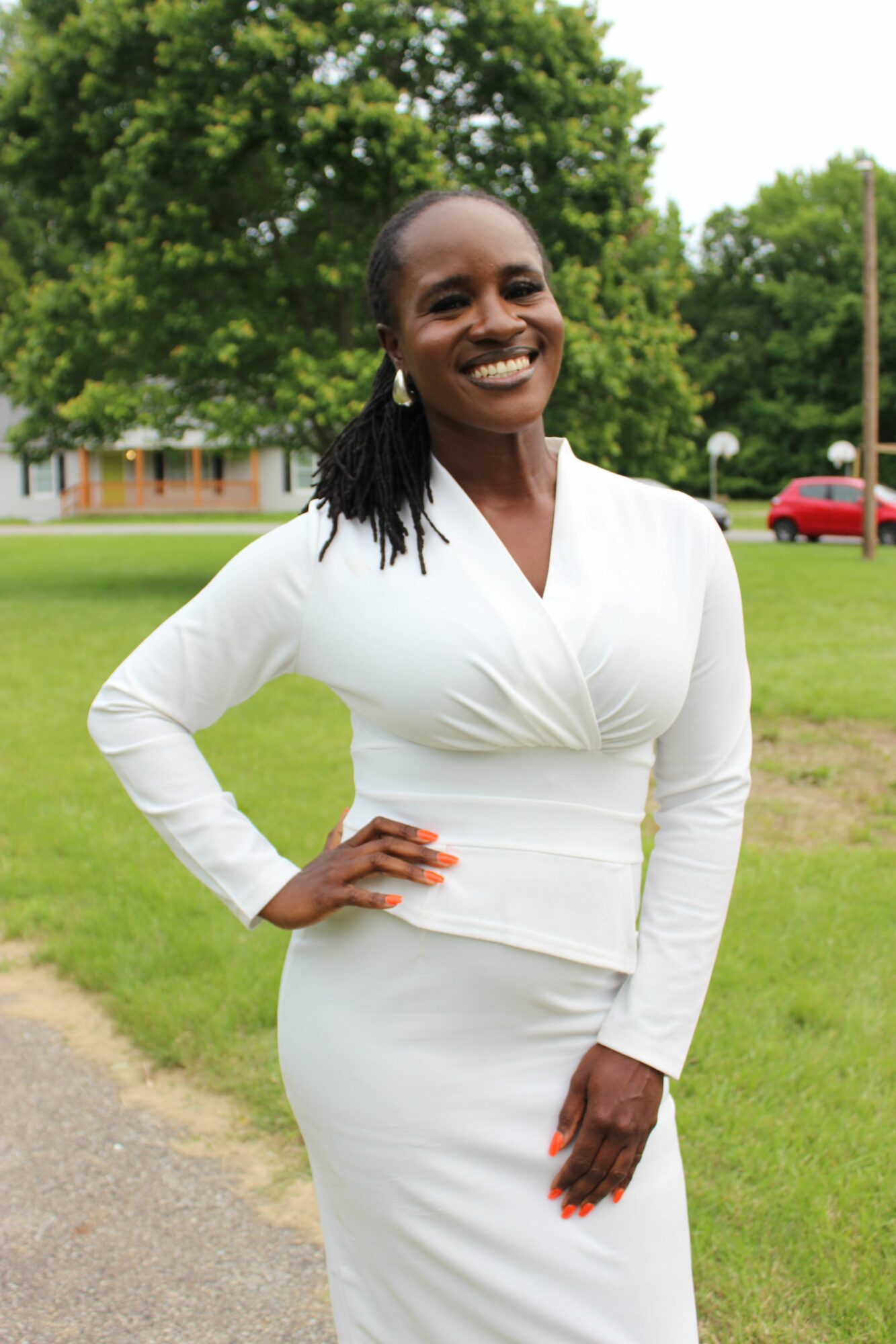 Smiling woman in white dress standing outdoors on grass with trees and a building in background.