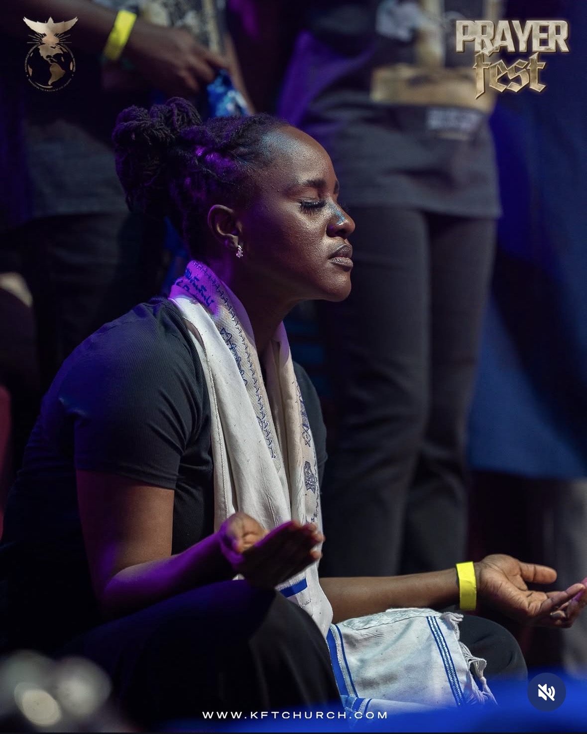 Woman with eyes closed, sitting with hands in prayer, wearing a scarf and black shirt, in a prayer setting.