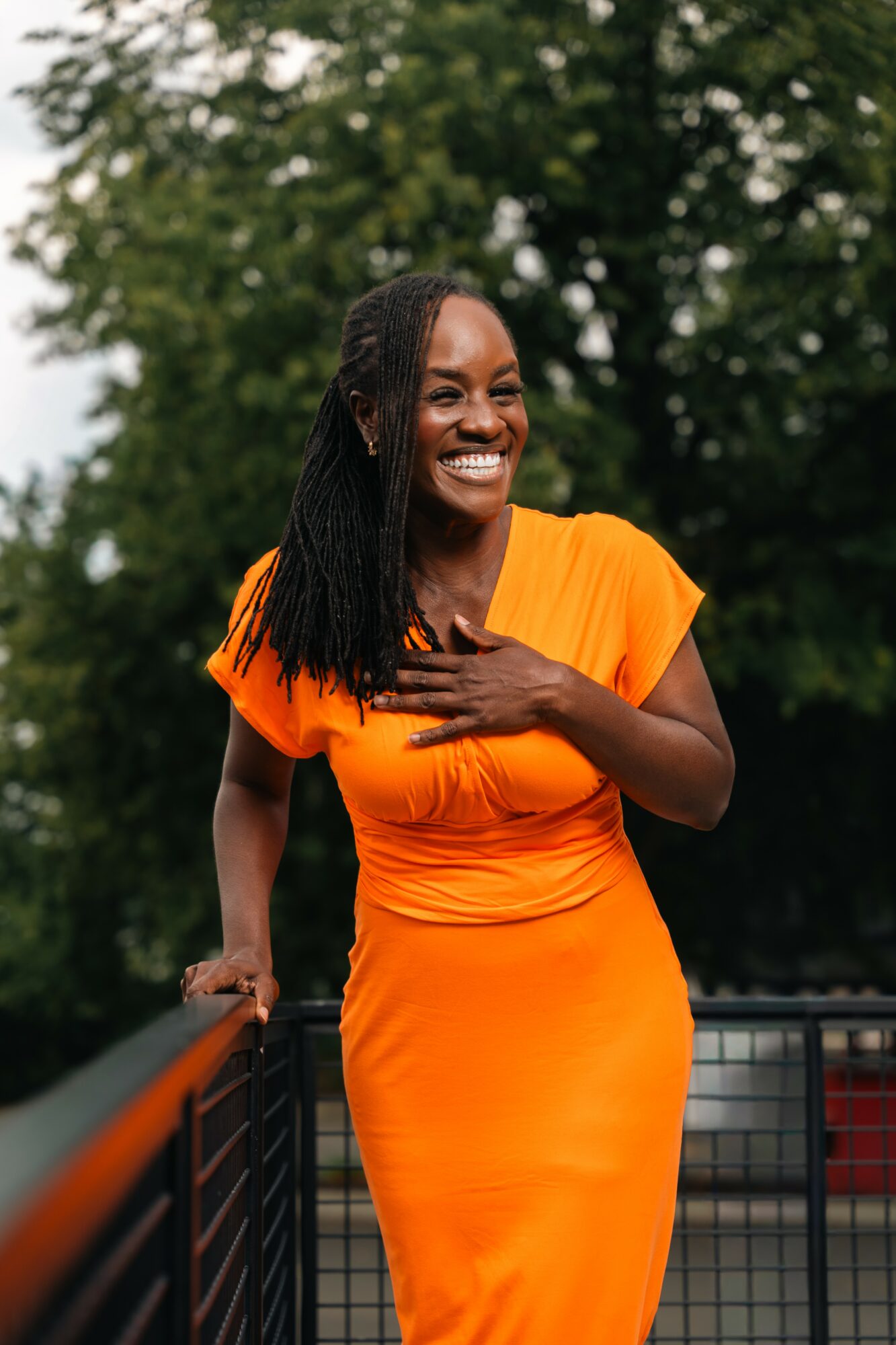 Woman in orange dress smiling outdoors, hand on chest, trees in background.
