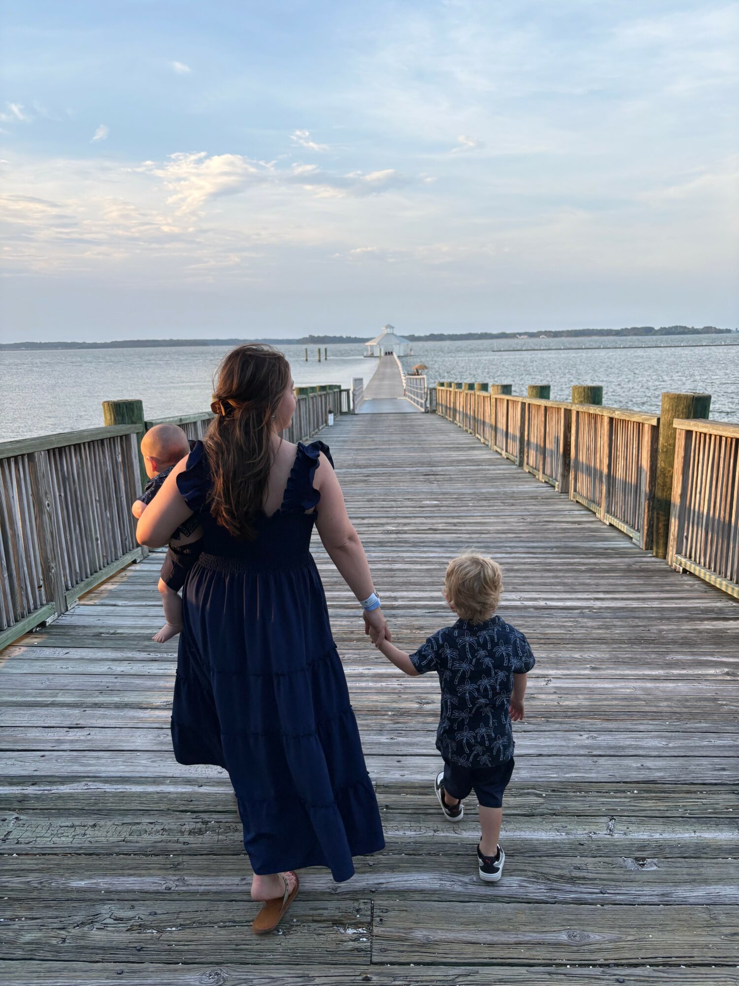 Woman holding child's hand walking on a wooden pier towards a lighthouse, with water and sky in the background.