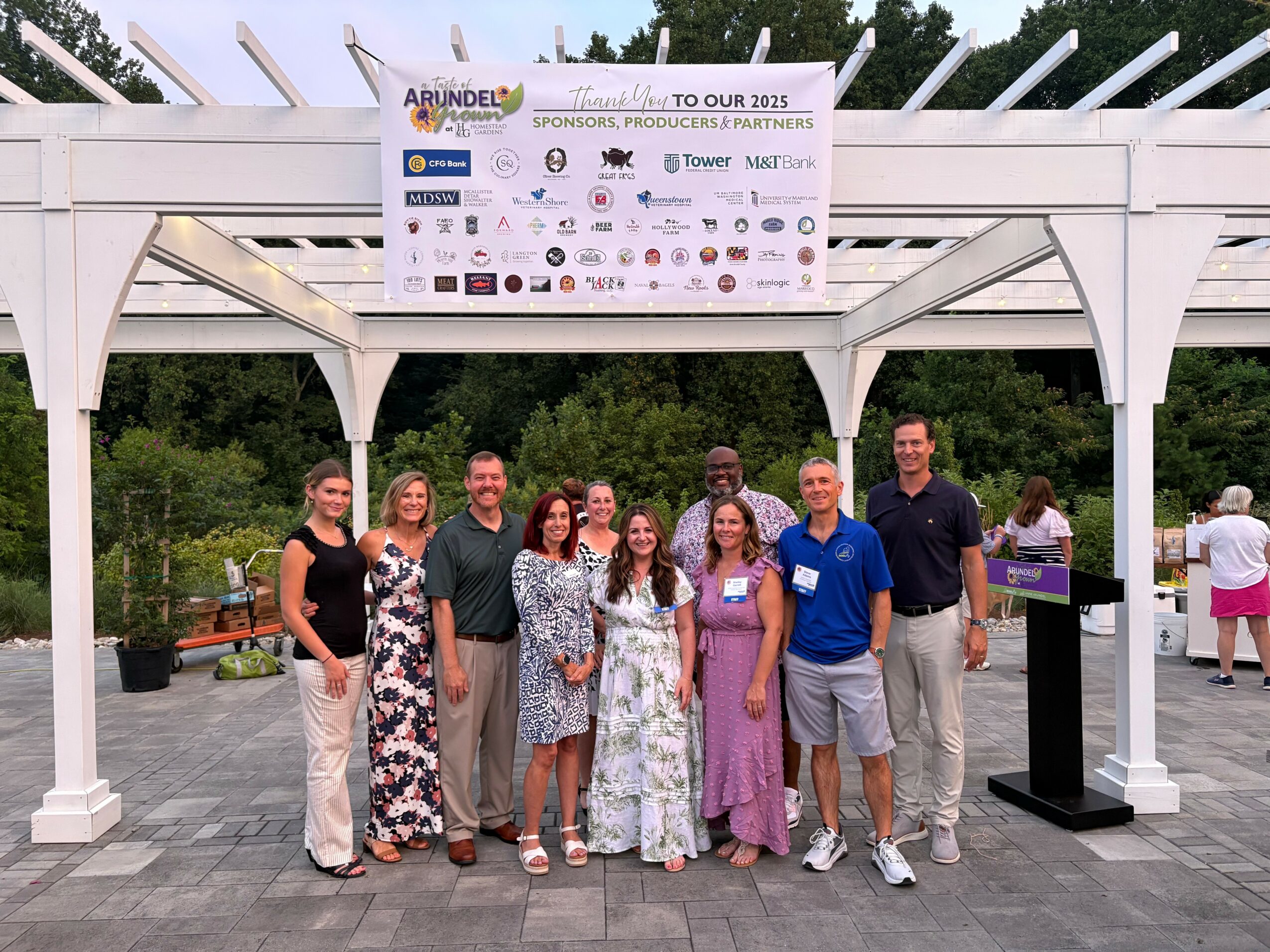 Group of nine people standing outdoors under a white pavilion with a banner above, trees in the background.