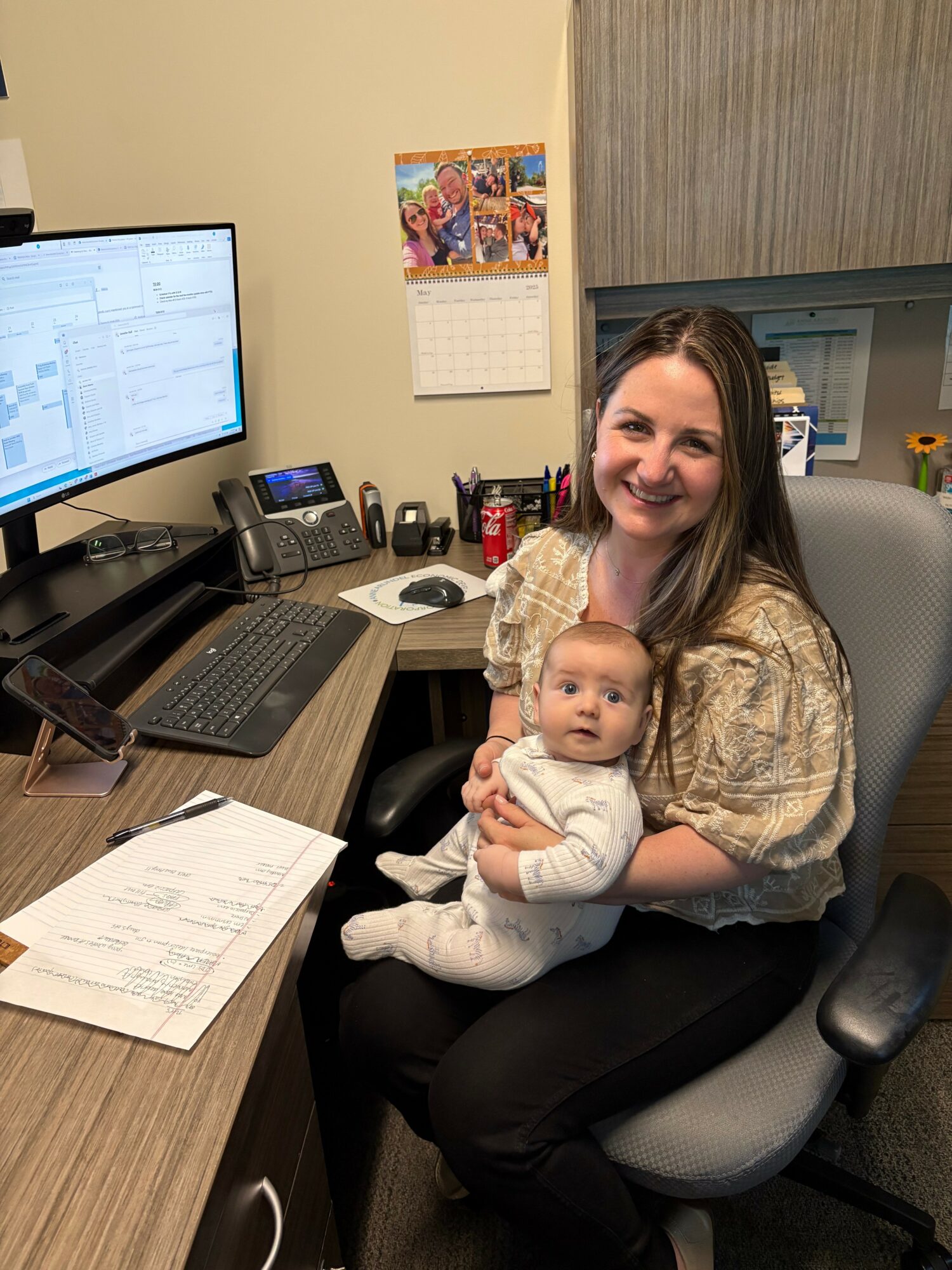 Woman sitting in an office chair holding a baby, smiling, with a desk, computer, and office supplies in the background.
