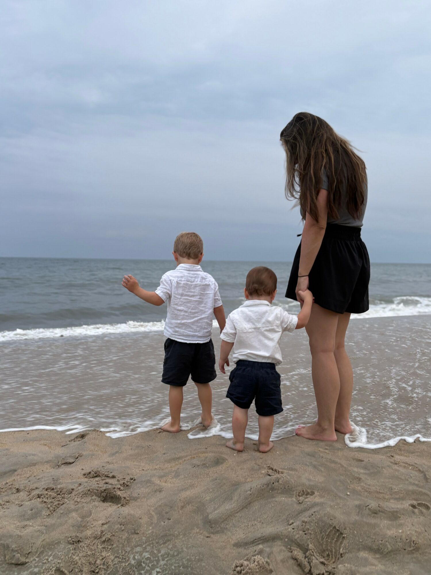 Woman and two children standing on the beach facing the ocean, with cloudy sky above.