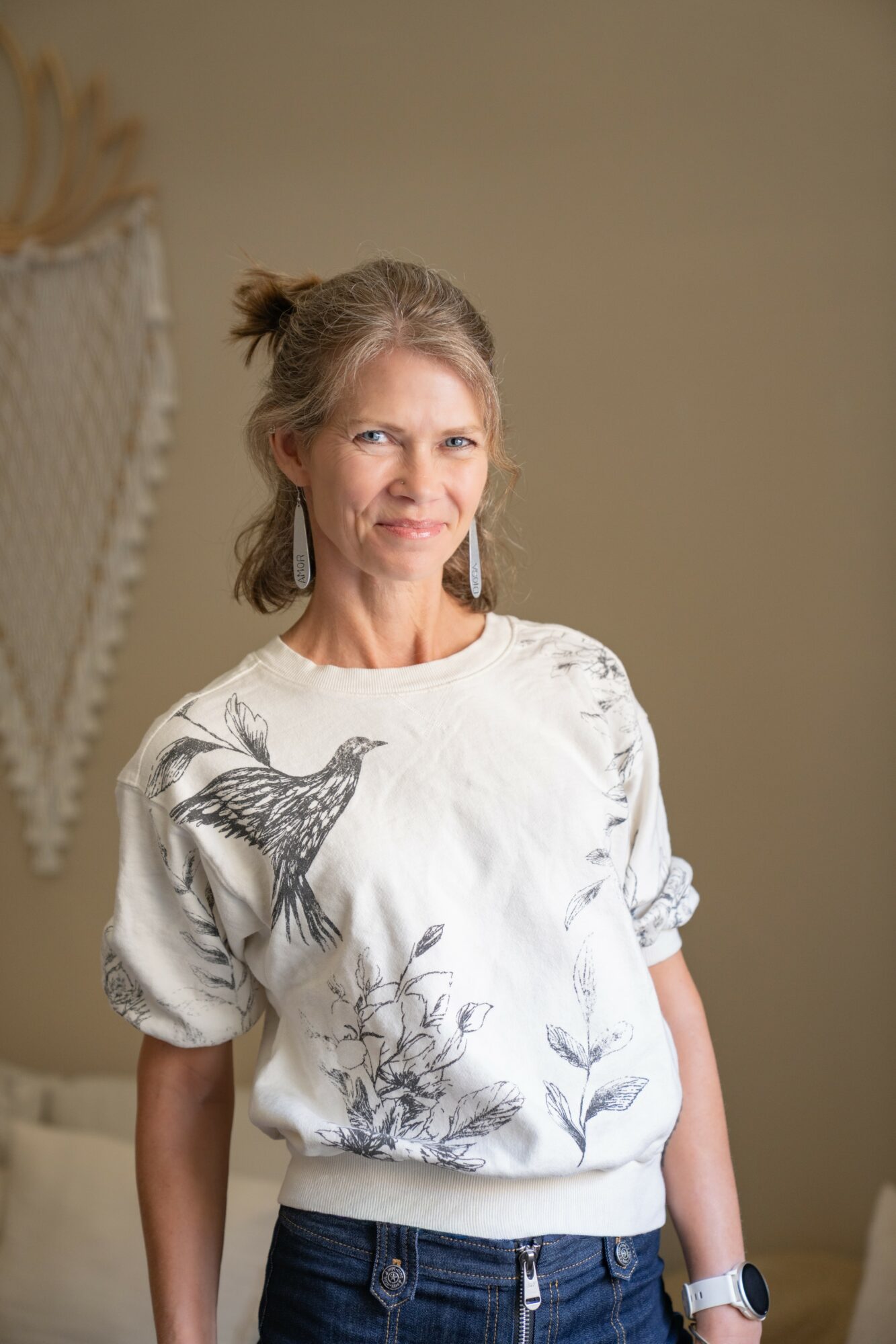 Woman with light brown hair in a bun, wearing a white sweatshirt with black floral and bird designs, standing indoors.