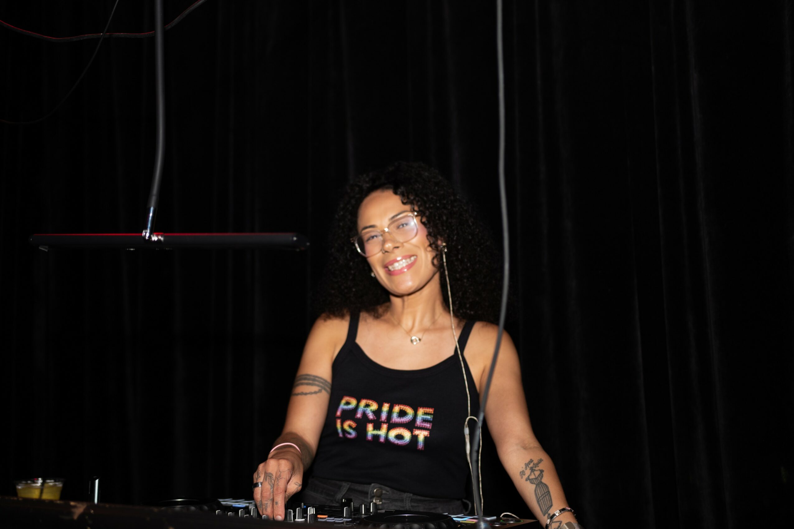 Smiling woman with curly hair and glasses stands behind DJ equipment on stage with black curtains.
