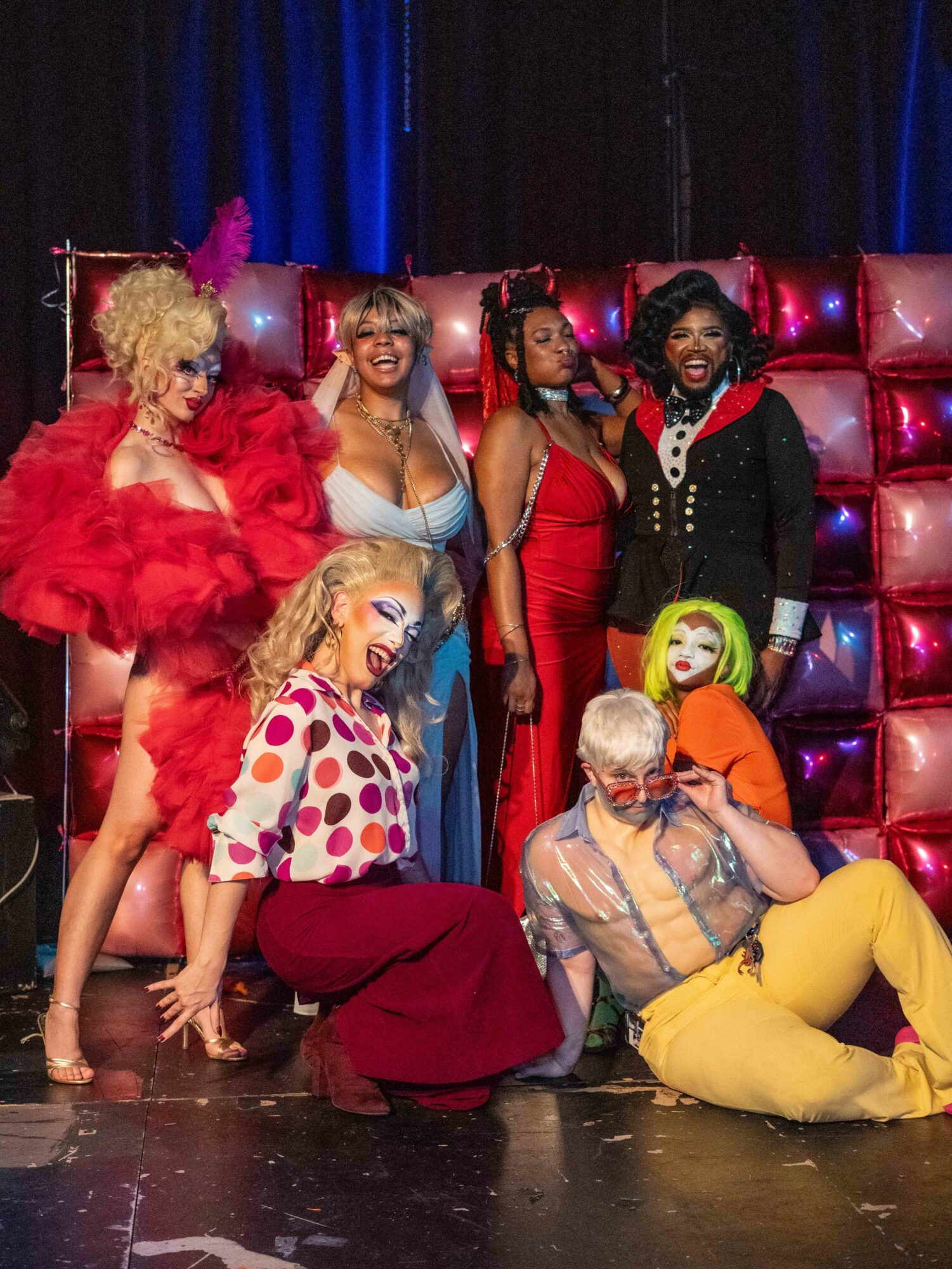 Group of seven performers in colorful costumes on stage with a shiny backdrop, some sitting and others standing, smiling.