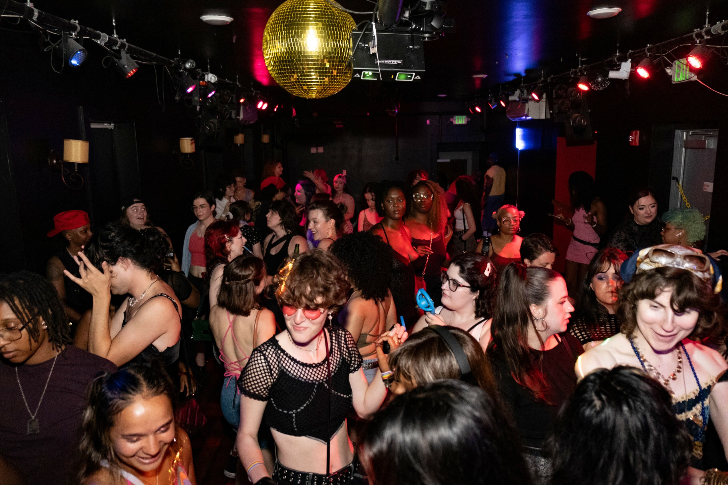 Crowd of people dancing and socializing in a dimly lit club with a large gold disco ball overhead.