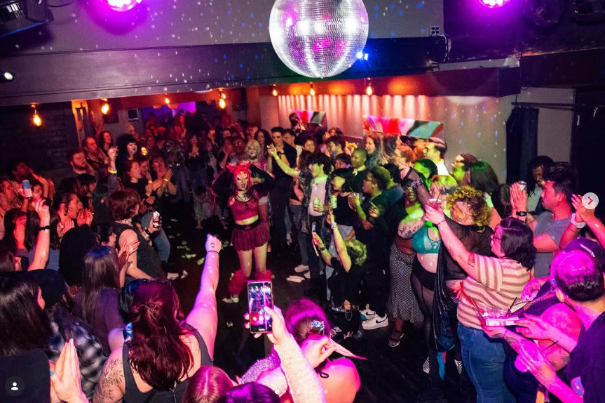 Crowd dancing and socializing in a nightclub with colorful lights and a disco ball overhead.